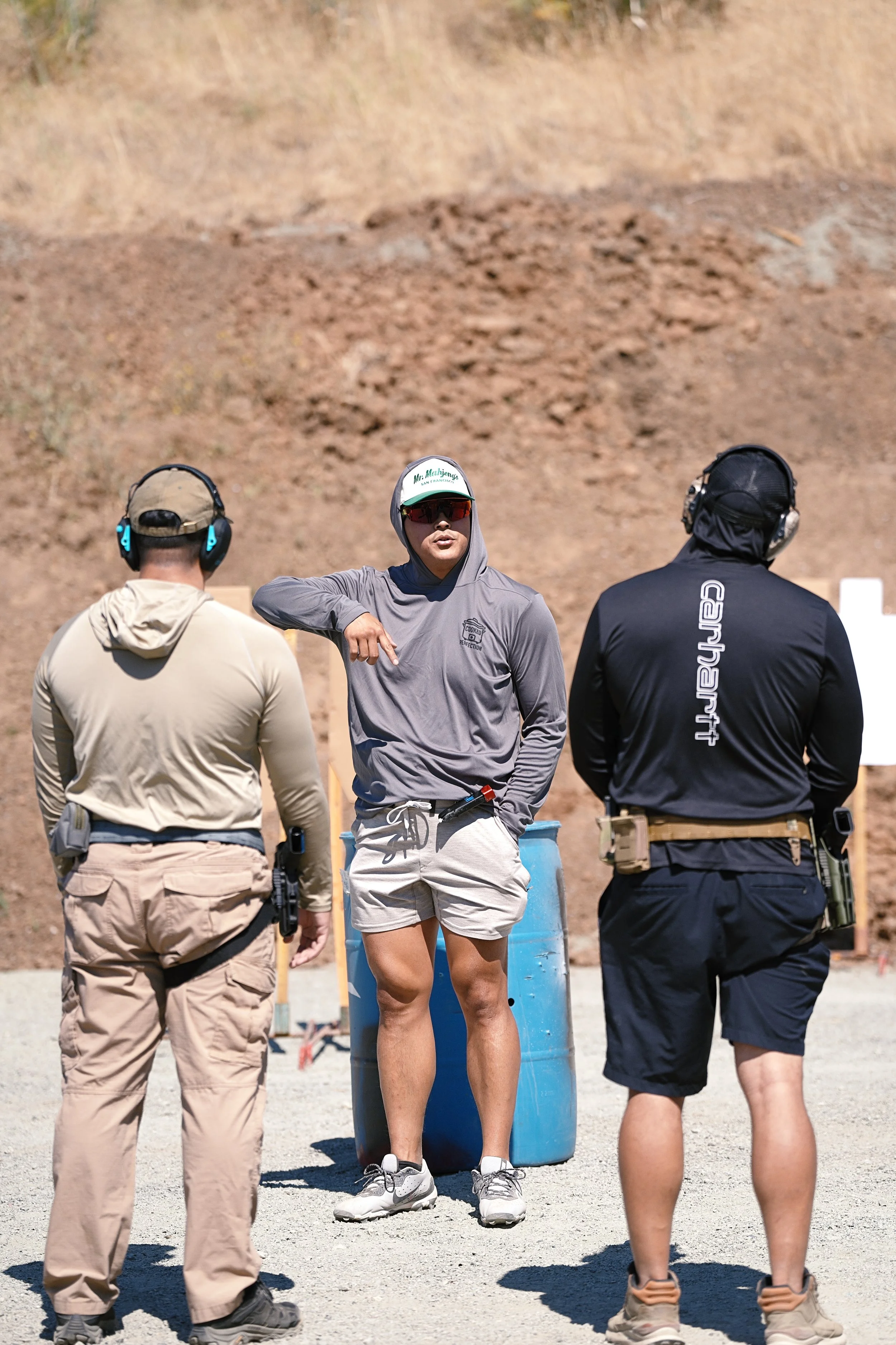 Three people at a shooting range, two men with hearing protection and a woman wearing sunglasses and a cap, standing in front of a blue barrel