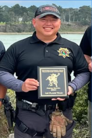 A smiling police officer in uniform holding a plaque and standing outdoors near water with trees in the background.