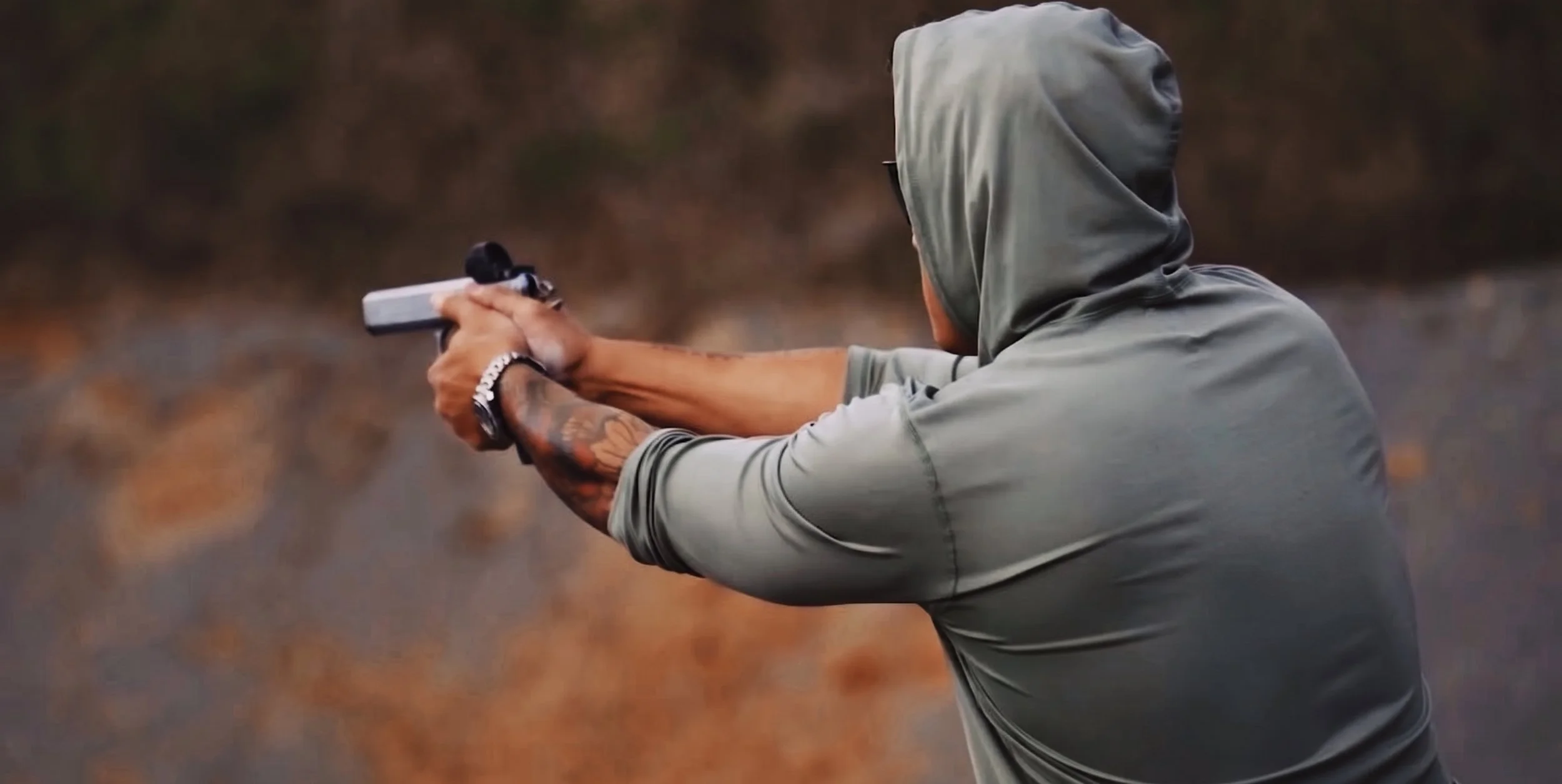 Person aiming a gun during outdoor activity, wearing a hoodie and tattoos on arms.