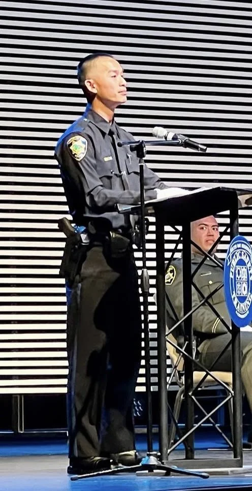 Police officer speaking at a podium with a microphone while another officer seated behind him, against a striped background.