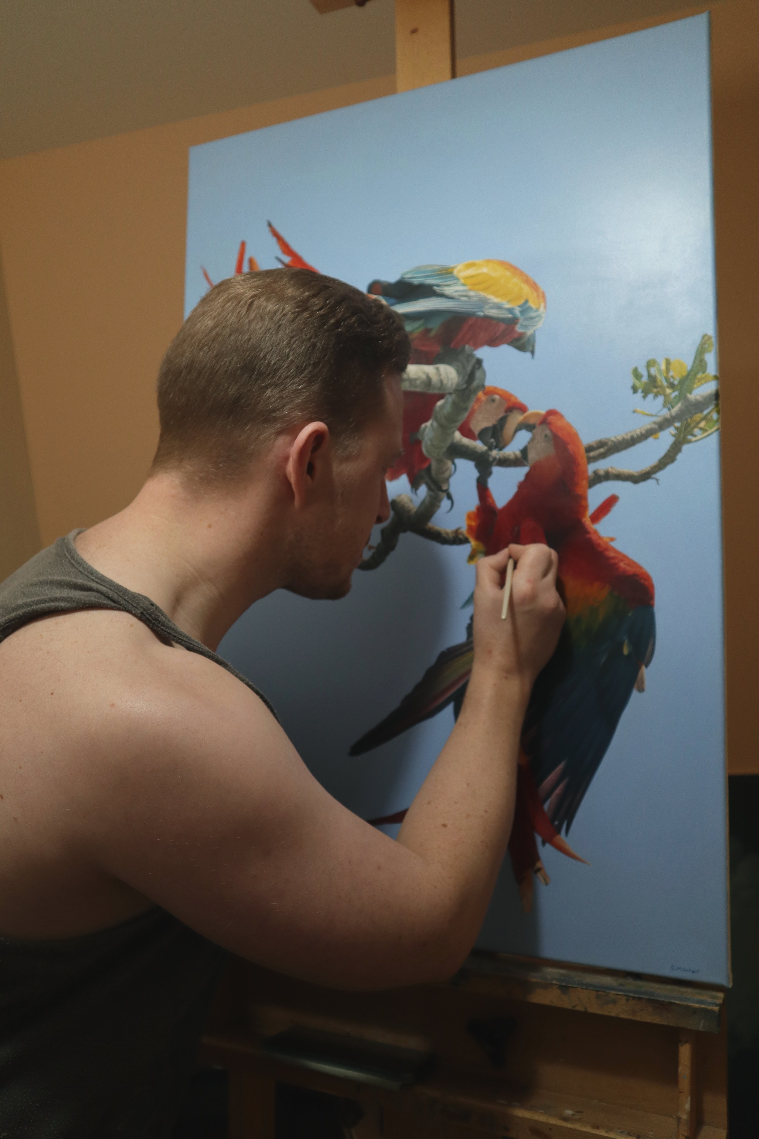 A man with short brown hair is painting a detailed picture of colorful parrots on a light blue canvas in an art studio.