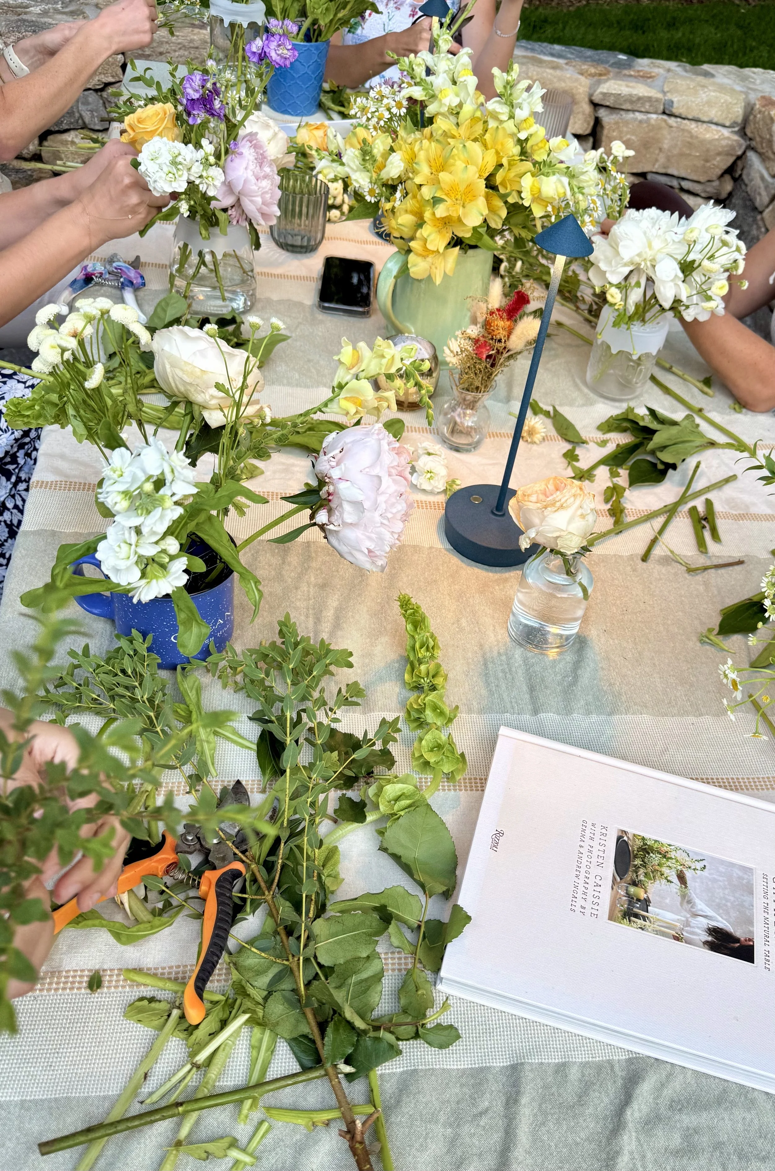 A table filled with various fresh flowers in vases, floral arranging tools, and a magazine about flowers, with people working on flower arrangements outdoors.