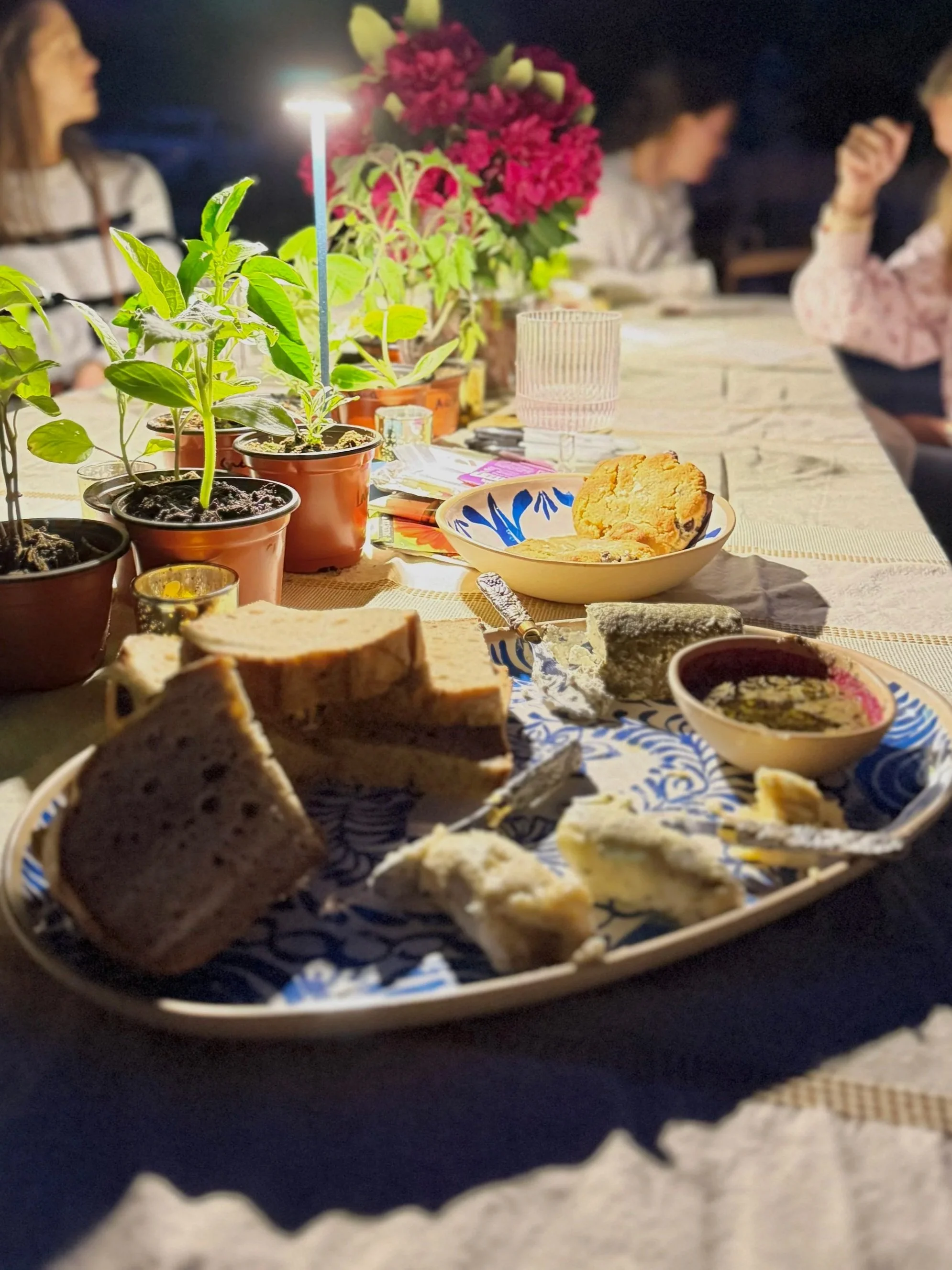 A table set with various foods, including sliced bread, a bowl of dip, and a plate of cookies, with potted plants and flowers in the background, and three women sitting and talking.