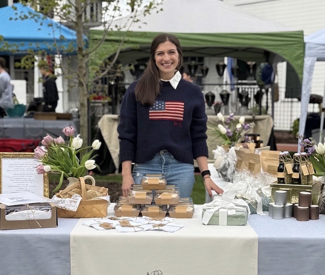 Women at a market stall with flowers and handmade products, smiling in front of green and white tents.