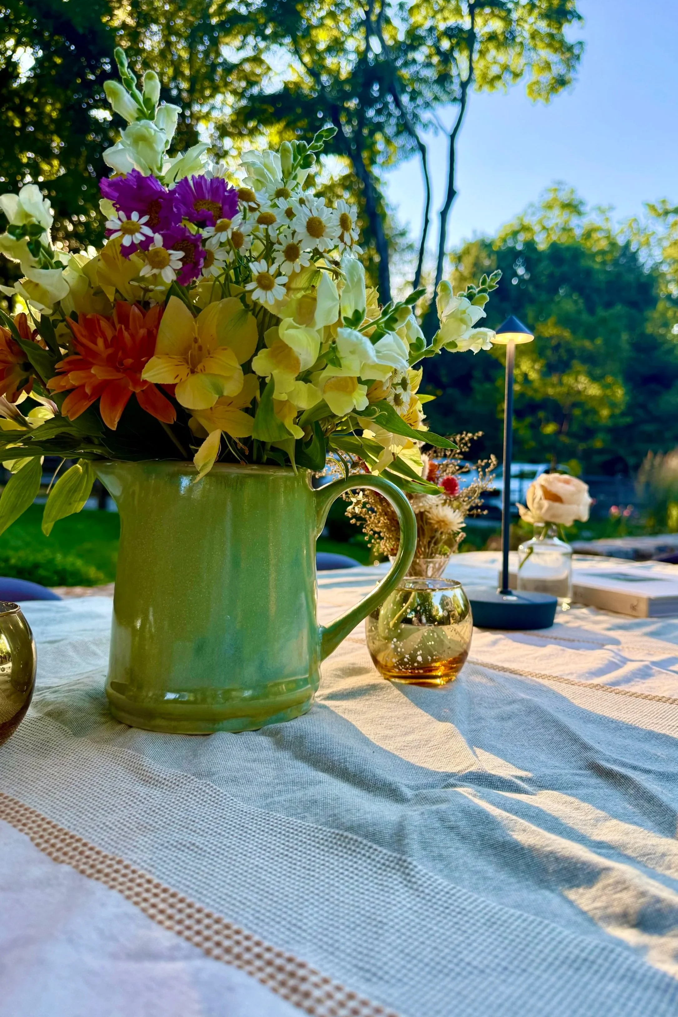 A green ceramic pitcher filled with a colorful bouquet of flowers, including daisies, gladiolas, marigolds, and other flowers, on a table with a white tablecloth. In the background, outdoor trees, a lamp, and additional small flower arrangements are visible, with sunlight filtering through the trees.