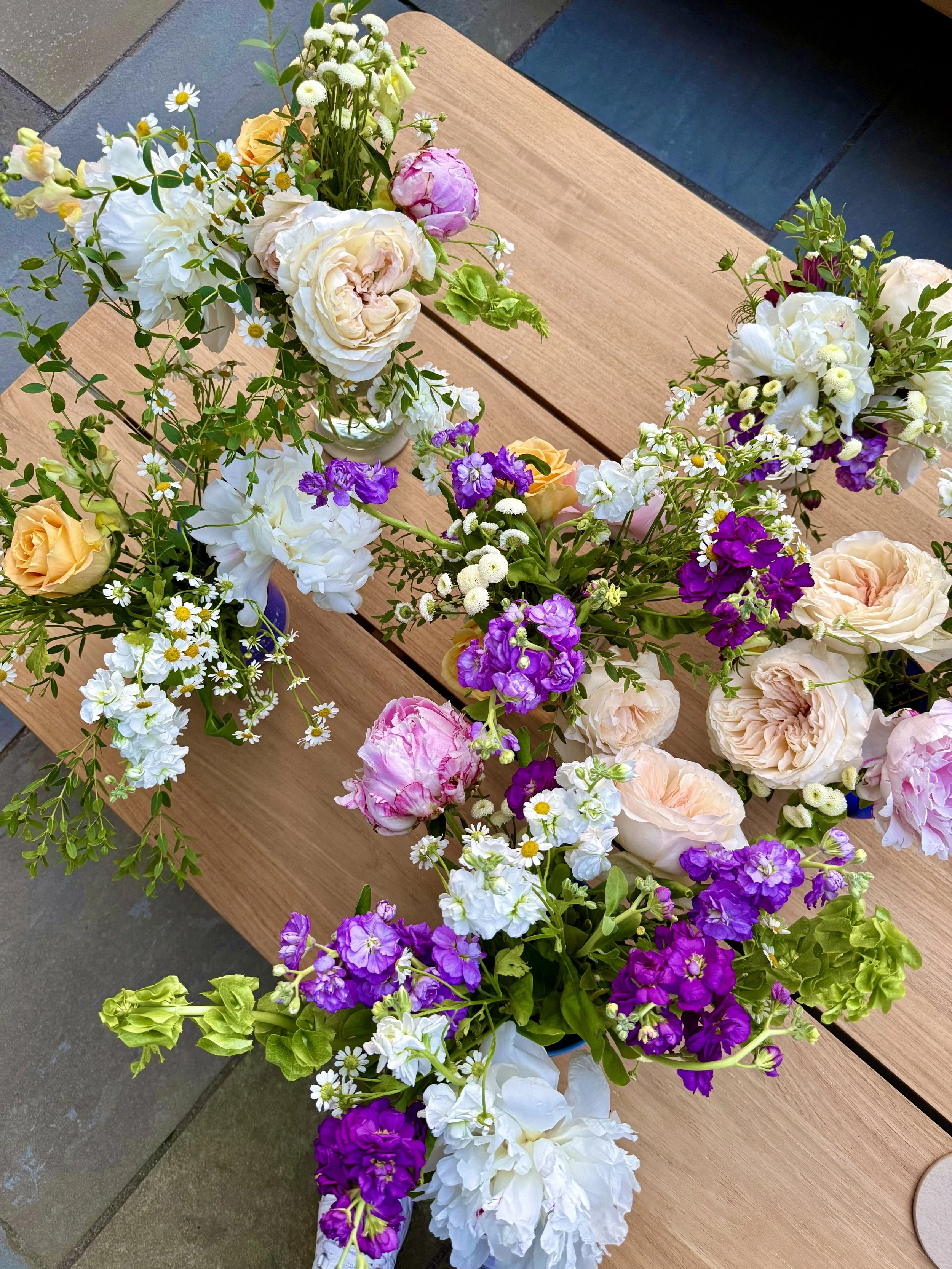 Colorful flower arrangements with roses, daisies, peonies, and other mixed flowers on a wooden table outside.
