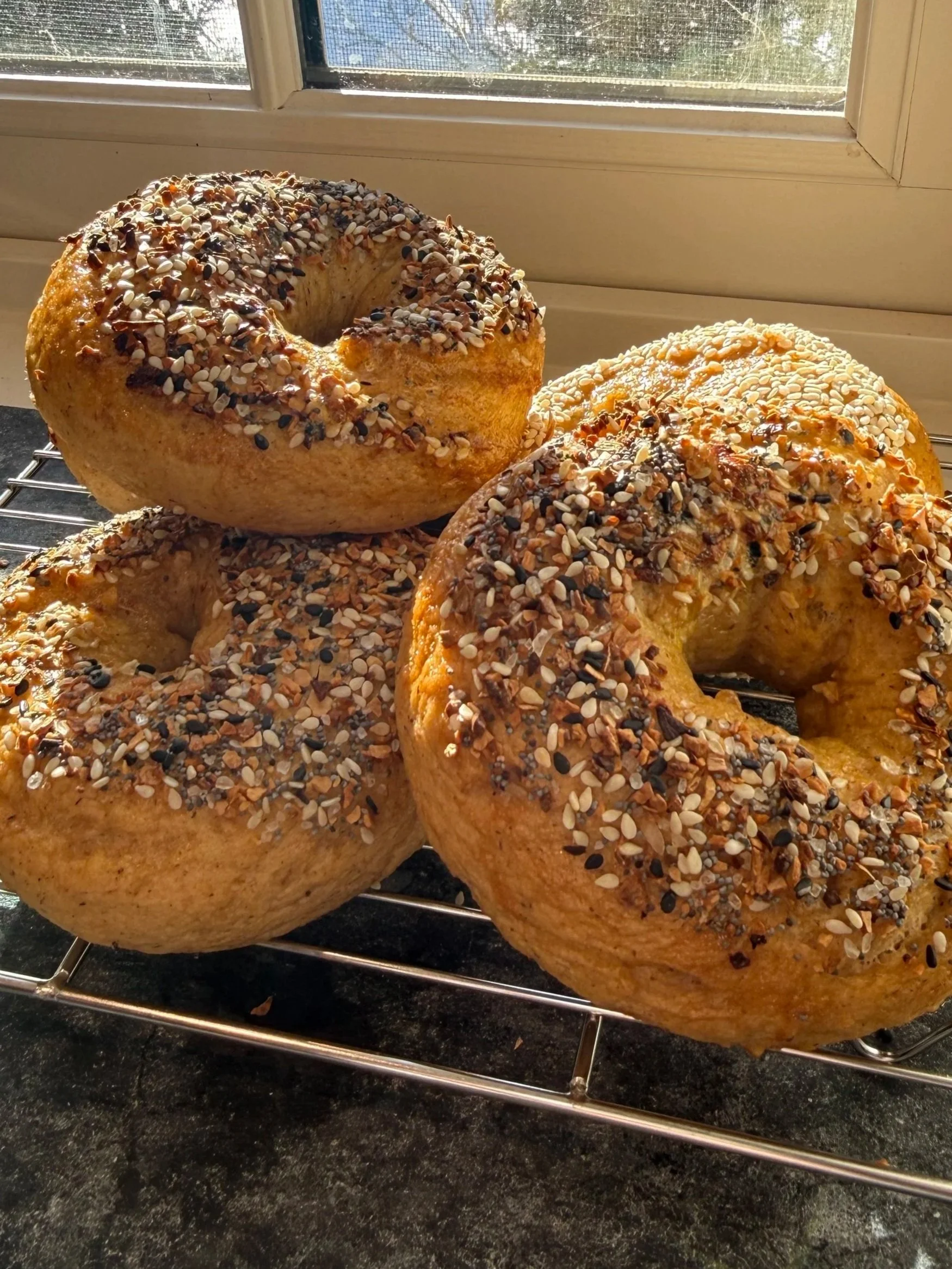 Three homemade bagels topped with mixed seeds and spices, cooling on a wire rack by a window.