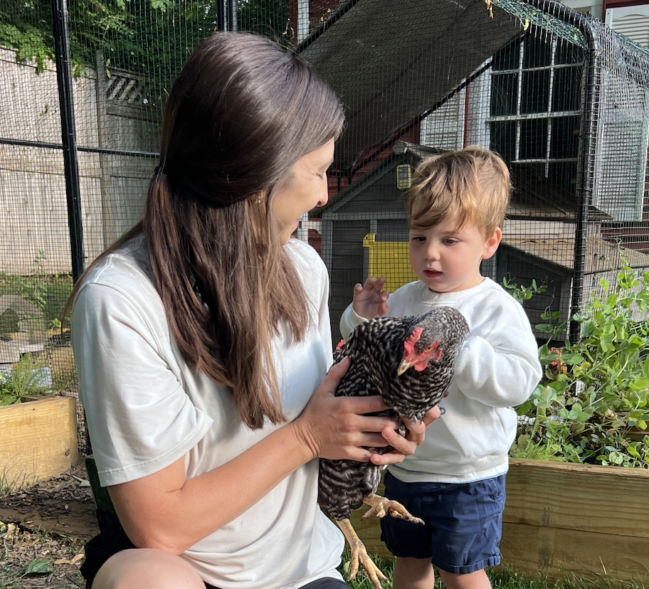 A woman and a young boy are outdoors, holding and looking at a chicken. The woman is smiling, and the boy appears curious. There is a chicken coop and plants in the background.