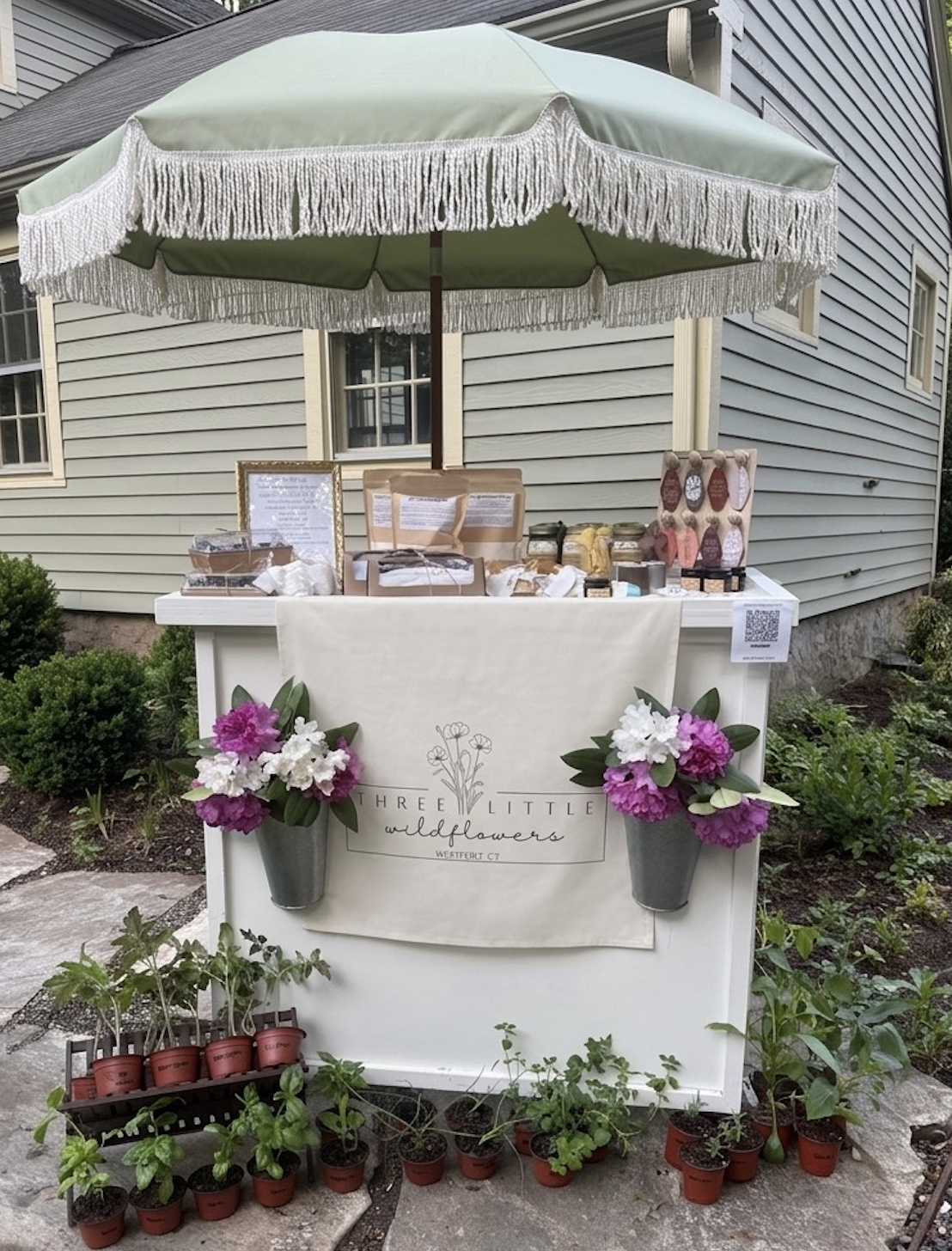 A flower stall with a green umbrella, flowers in pots, a white sign reading 'Three Little Wildflowers,' and various small plants and flowers on a white table in front of a house.