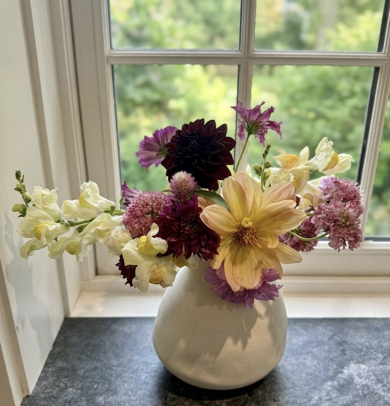 A vase with a colorful bouquet of flowers on a windowsill, with a blurred outdoor scene visible through the window.