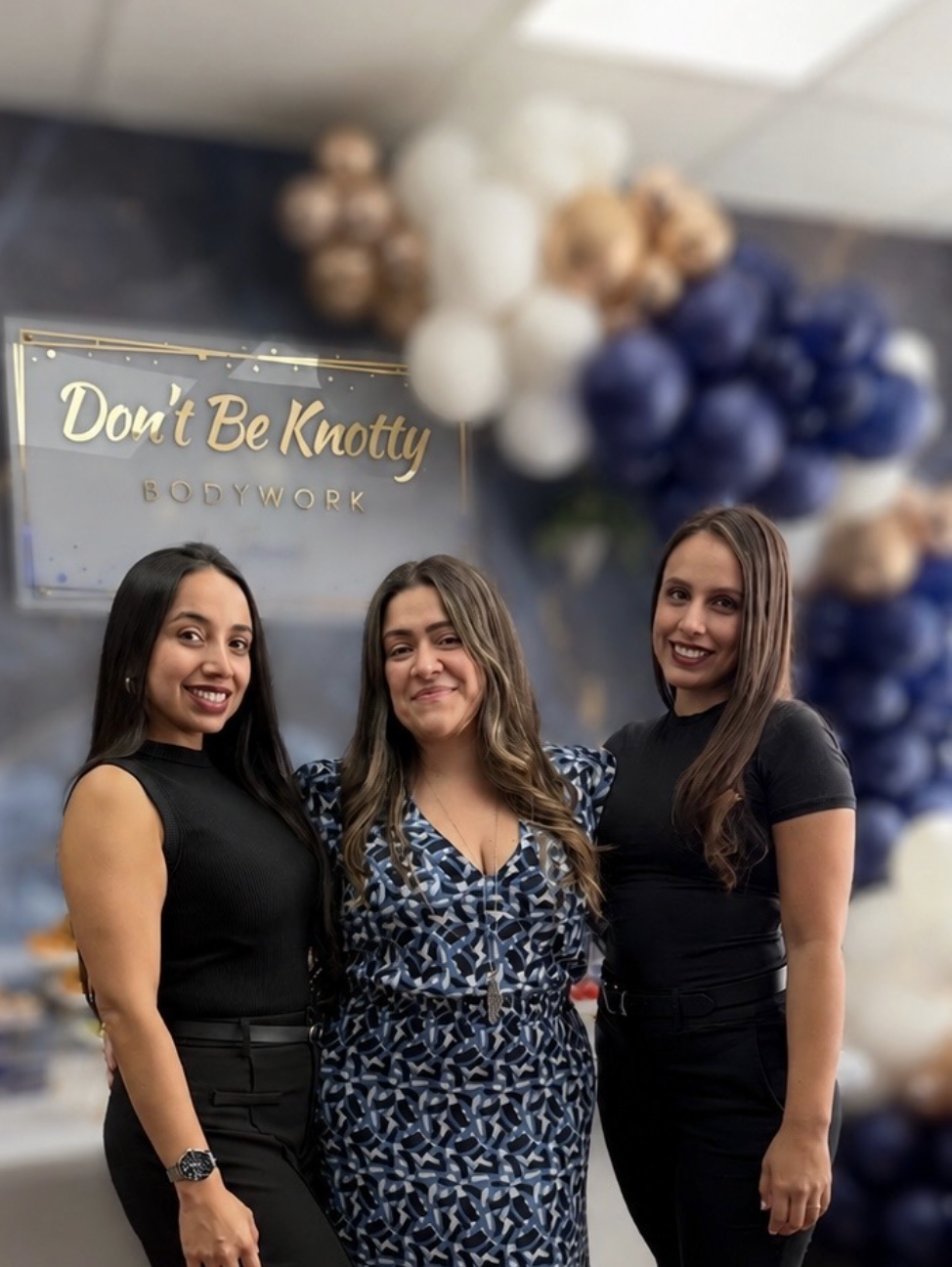 Three women standing inside a business named 'Don't Be Knotty Bodywork,' with balloon decorations in white, gold, and navy blue in the background.