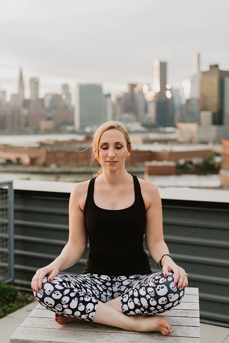 A woman practicing yoga outdoors on a rooftop with a city skyline in the background.