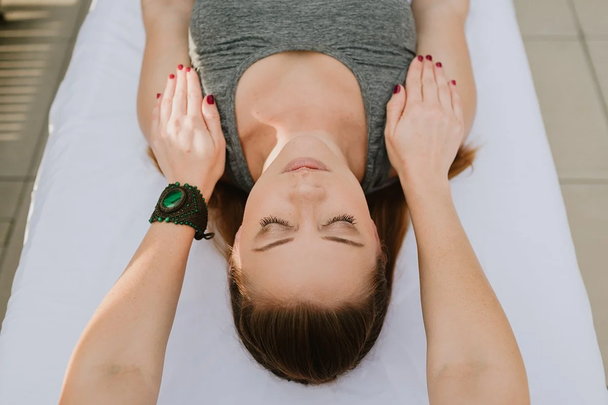 A woman with closed eyes lying on her back on a white massage table, receiving a massage or therapy, with her hands resting on her chest. She has long brown hair, red nail polish, and is wearing a gray sleeveless top. A green bracelet with beads is on her left wrist.
