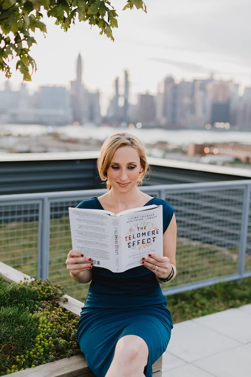 A woman in a dark blue dress sitting on a bench outdoors, reading a book titled 'The Telomere Effect' against a city skyline with river and buildings in the background, and some green leaves at the top left corner.