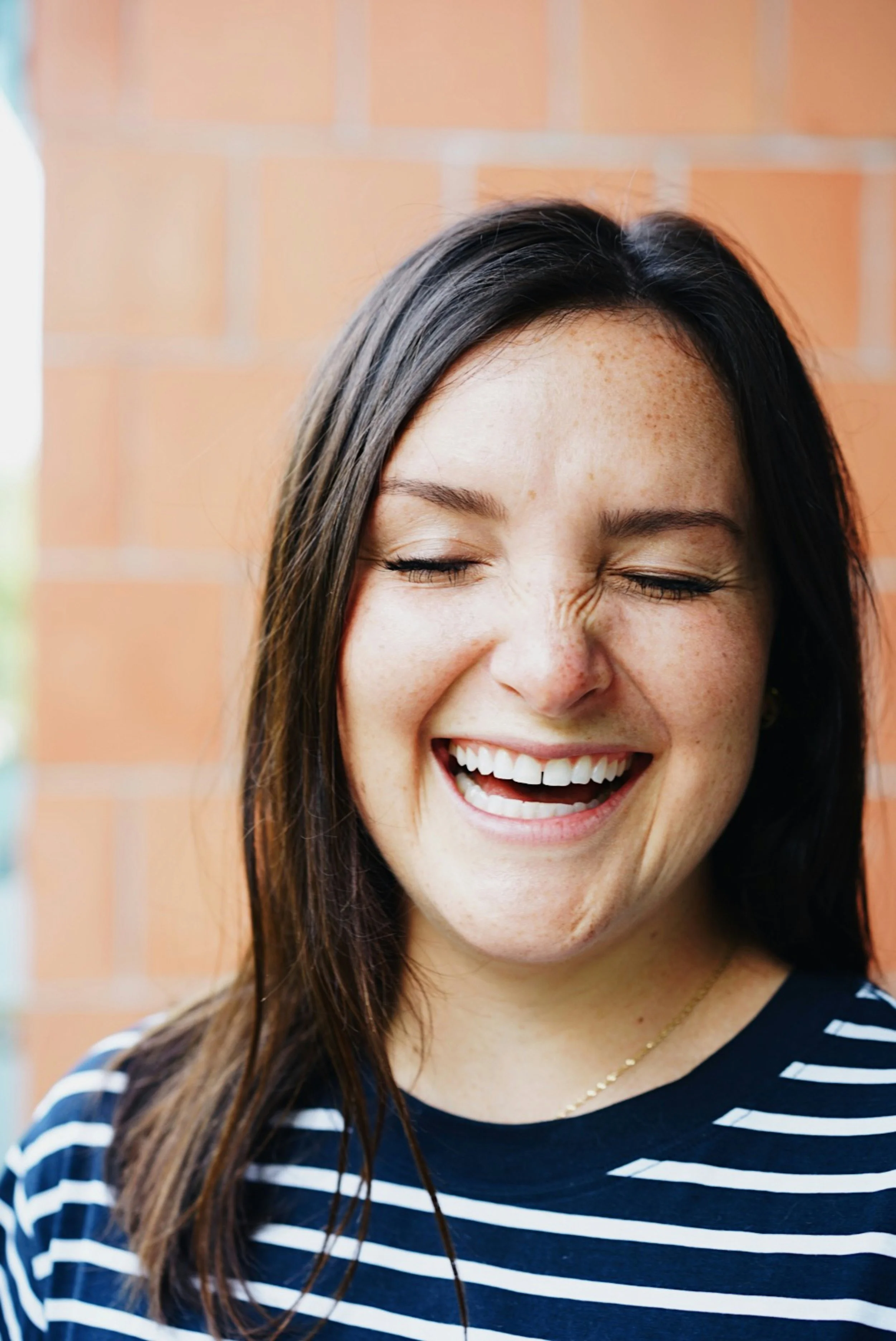 A young woman with long dark hair, freckles, and a broad smile showing her teeth, wearing a navy blue and white striped shirt, standing in front of a brick wall.