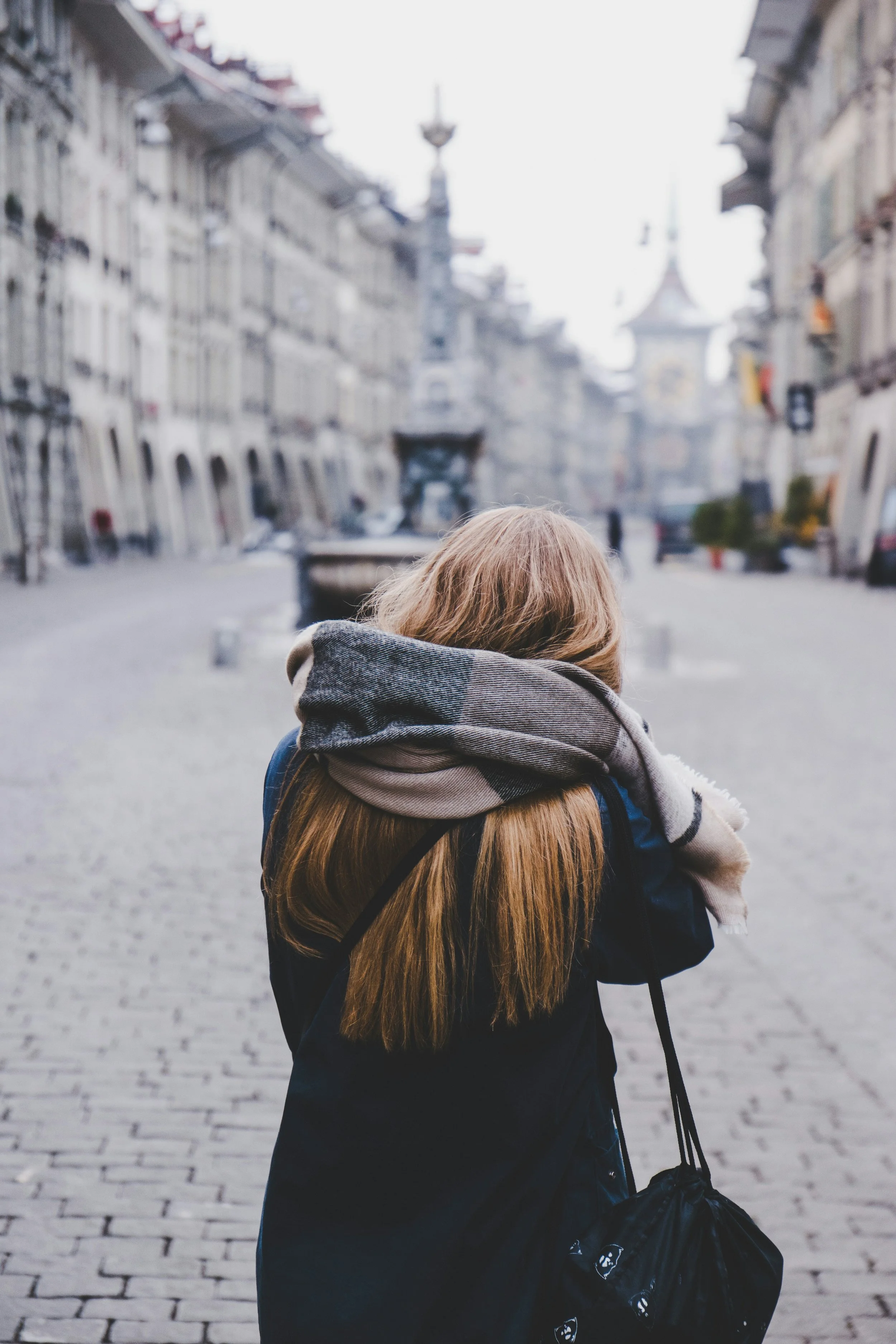 A person with long, light brown hair wearing a dark jacket and a gray and black plaid scarf, carrying a black bag with animal face prints, standing on a cobblestone street in a historic European city with old buildings and a church steeple in the background.