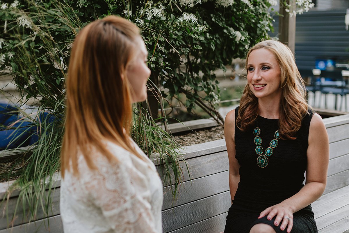 Two women sitting on a wooden bench outdoors, engaged in conversation, with greenery and flowers behind them.