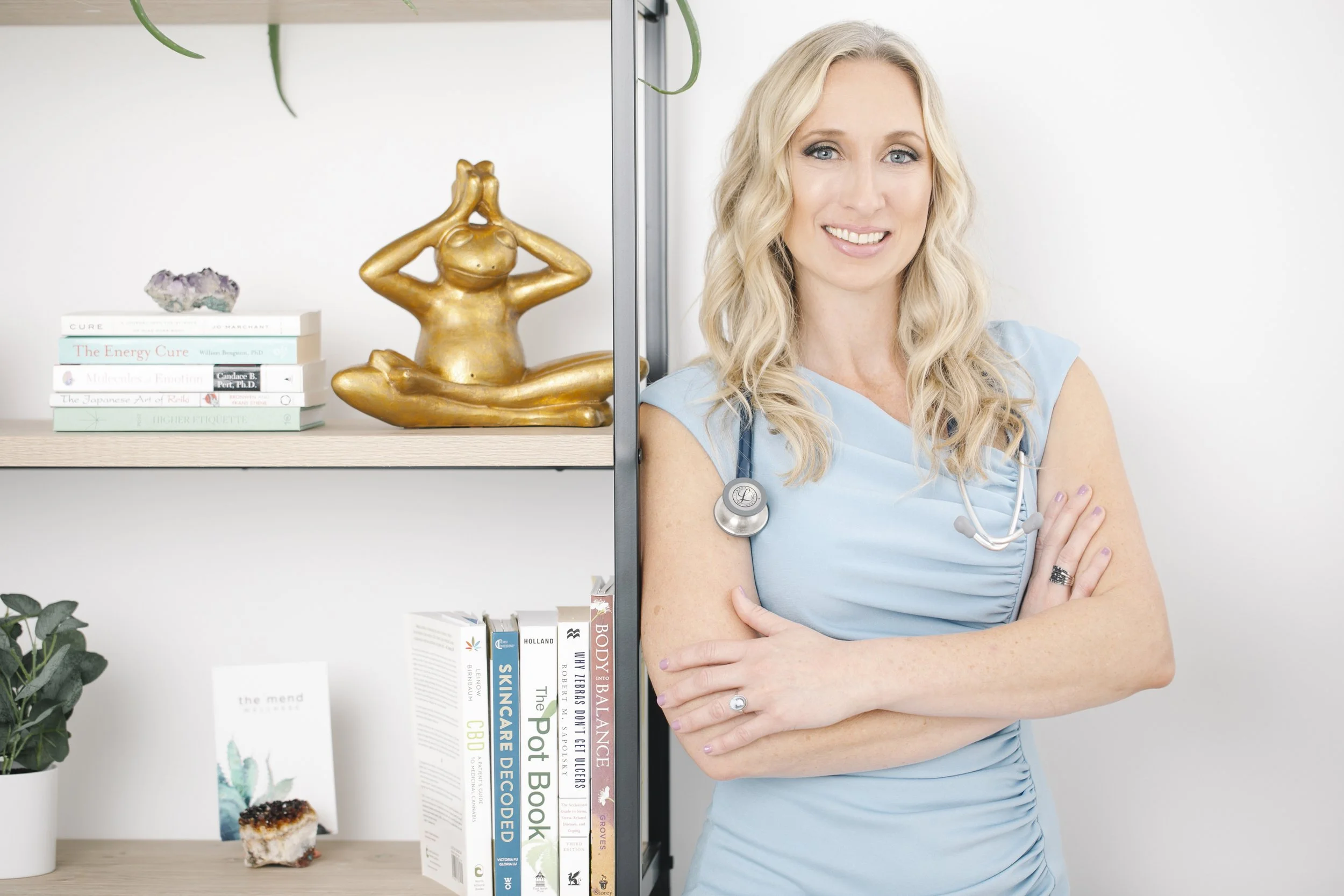 A woman in a light blue top with a stethoscope around her neck standing with her arms crossed in front of a white wall. There is a bookshelf with books, a gold-colored yoga statue, and some decorative items on the bookshelf.