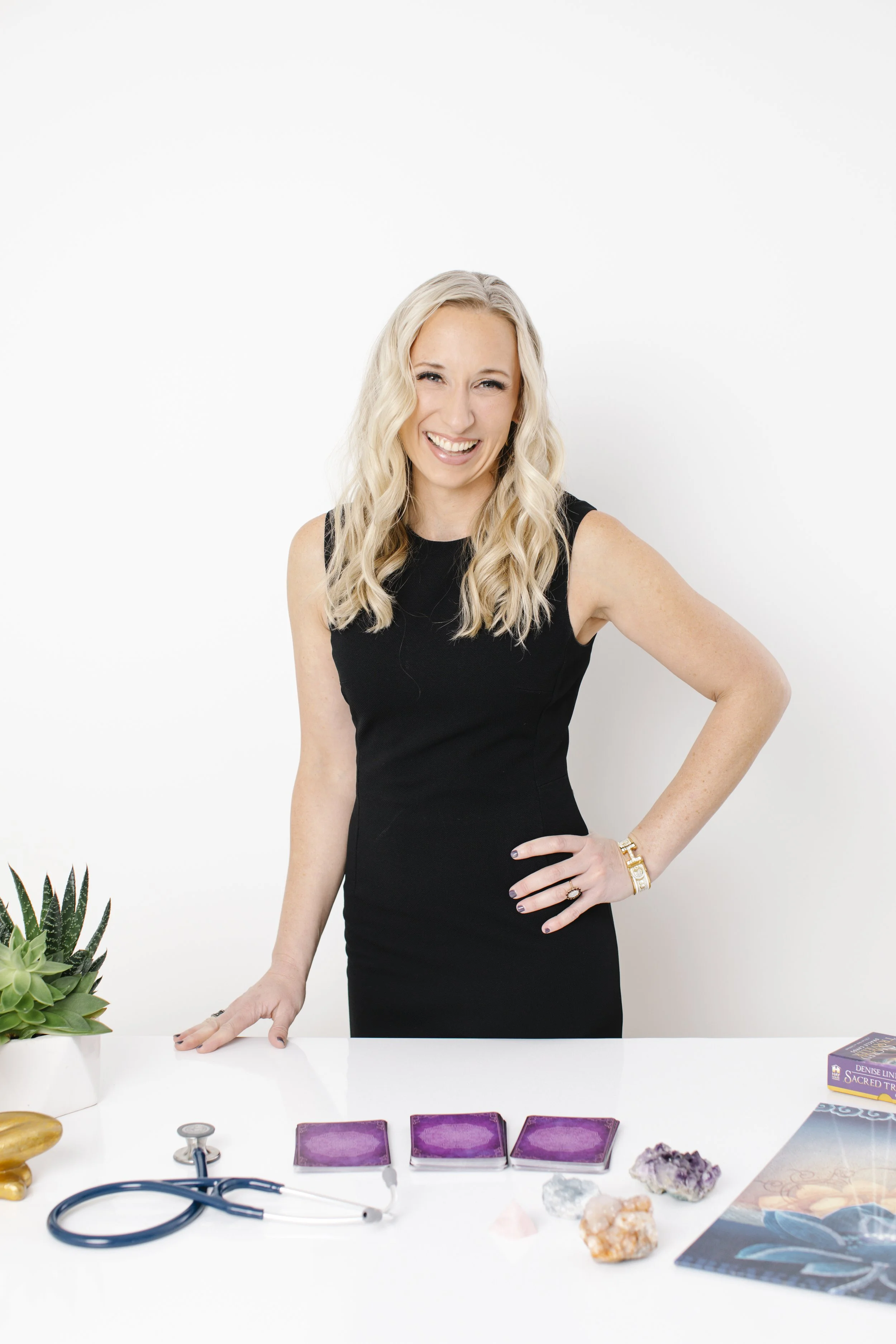 A woman with long, wavy blonde hair wearing a black sleeveless dress, standing behind a white table, smiling and slightly tilting her head. On the table are various items including a potted succulent plant, purple tarot cards, a stethoscope, crystals, and books. The background is plain white.