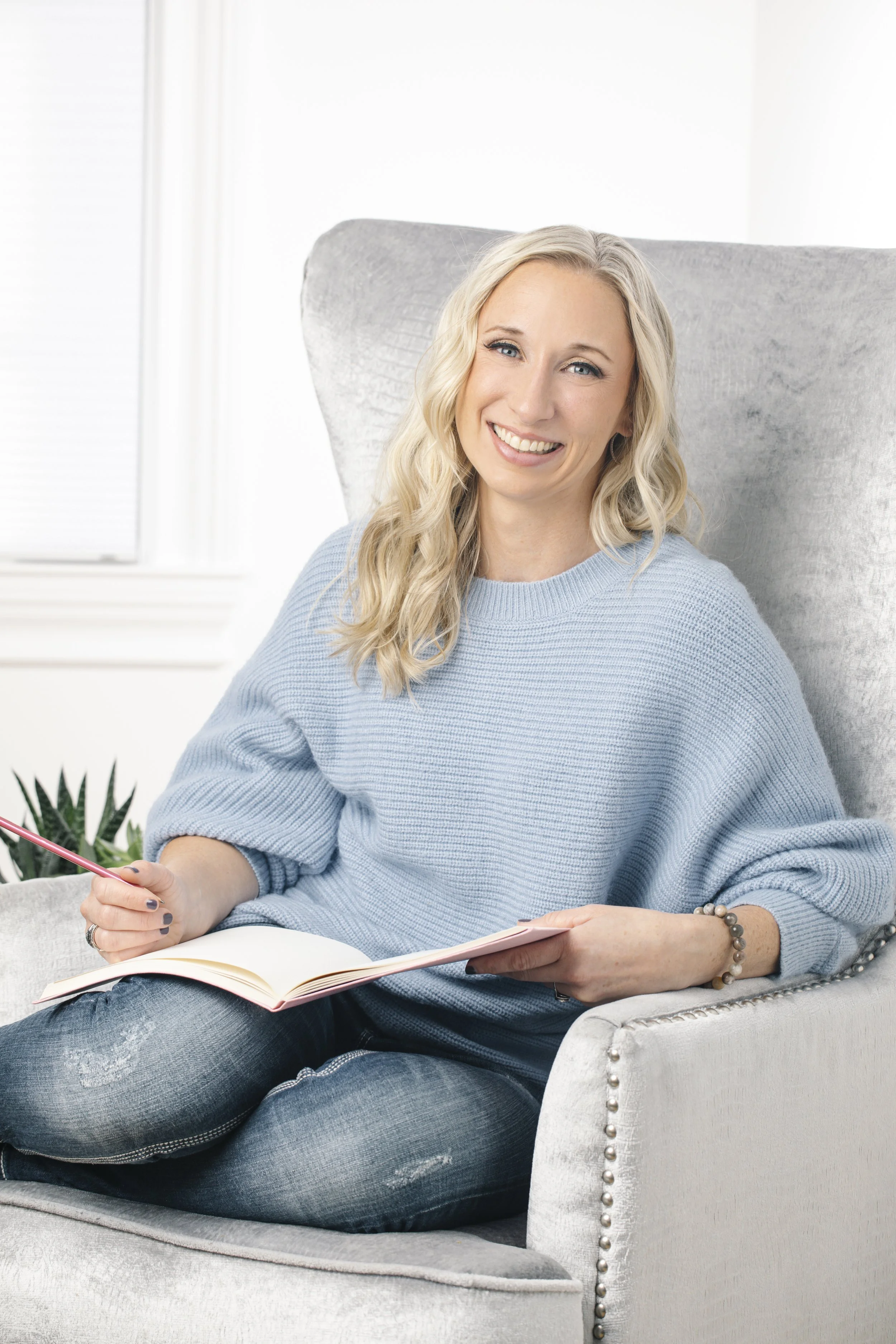 A woman with blonde curly hair smiling while sitting on a light gray armchair, holding a pink pen and an open notebook, dressed in a blue sweater and distressed jeans, in a bright room with white walls and a window.