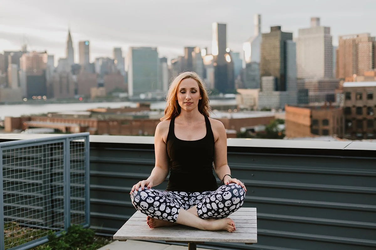 A woman practicing yoga in meditative pose on a rooftop with city skyline in the background.