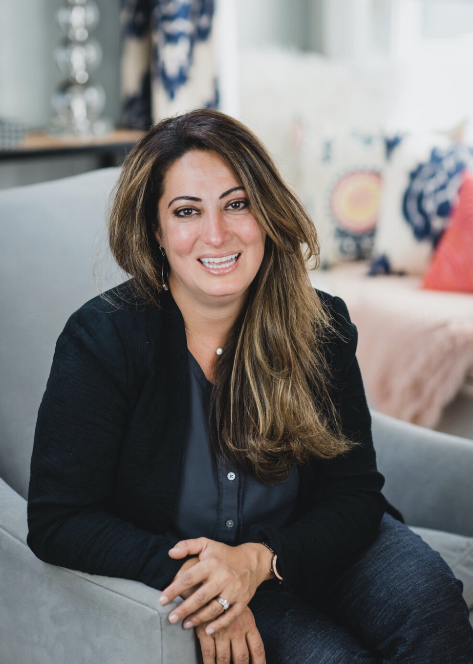 A woman with long brown hair, smiling and wearing a black blazer and dark shirt, sitting on a light gray sofa in a cozy living room with colorful pillows and decor.