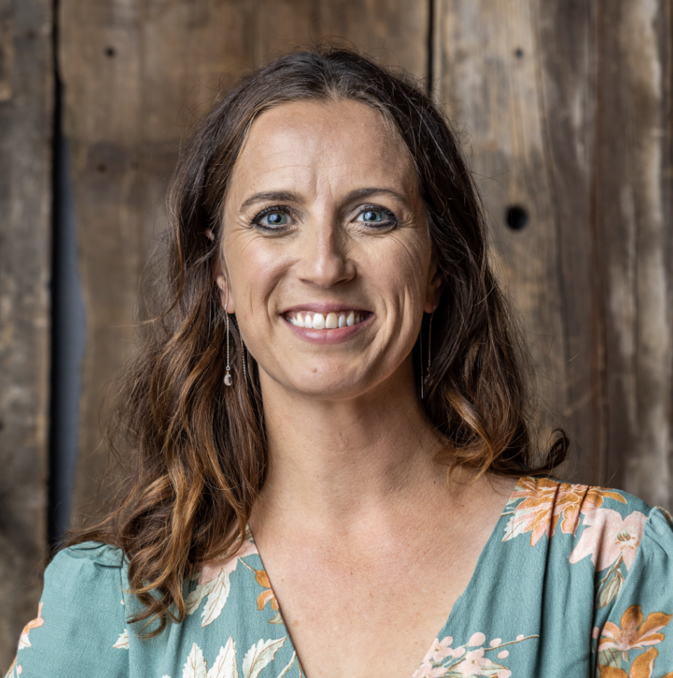 Portrait of a woman with long brown hair, blue eyes, wearing earrings and a floral patterned dress, standing in front of a wooden background.