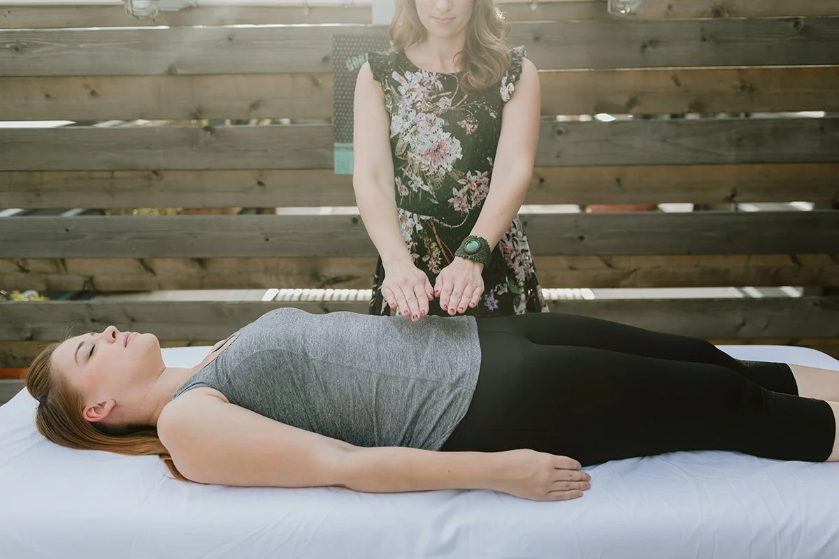 Woman lying on her back on a massage table with eyes closed, receiving a hand massage from a woman standing next to her, outdoors with a wooden fence in the background.
