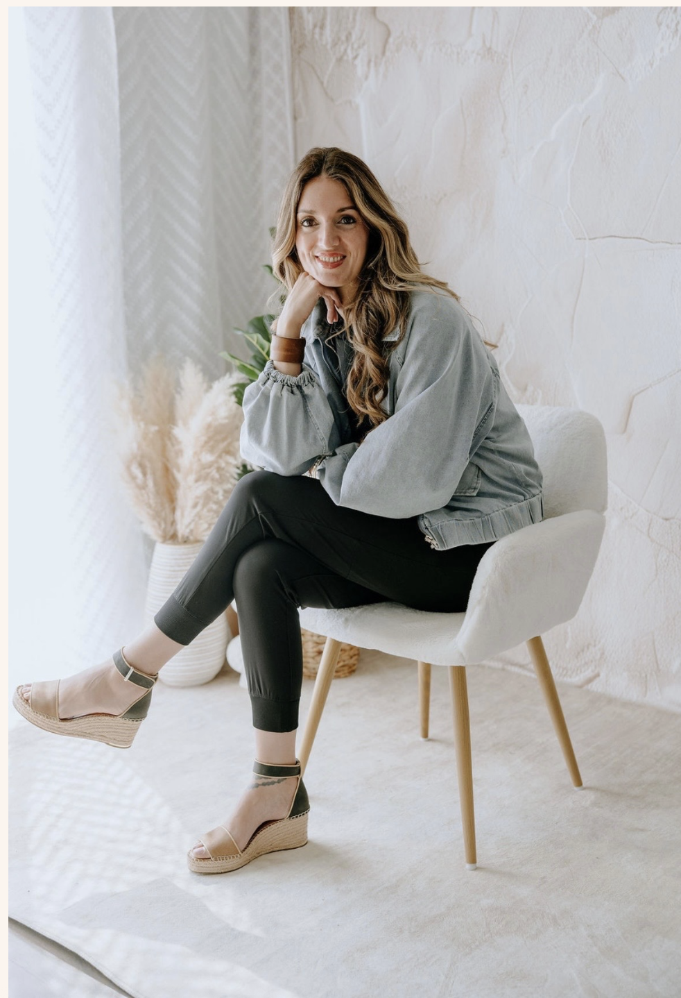 A woman with long wavy hair, smiling, sitting on a white chair with wooden legs in a bright room with textured walls and a neutral-toned carpet. She is wearing a light denim jacket, black pants, and beige and black wedge sandals.