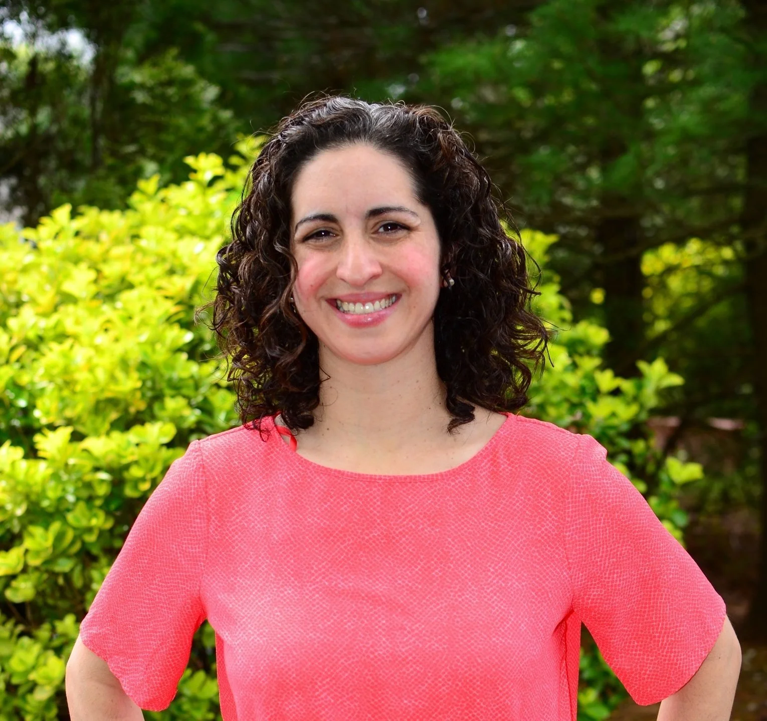 A woman with dark curly hair wearing a coral pink top, standing outdoors with green foliage in the background.