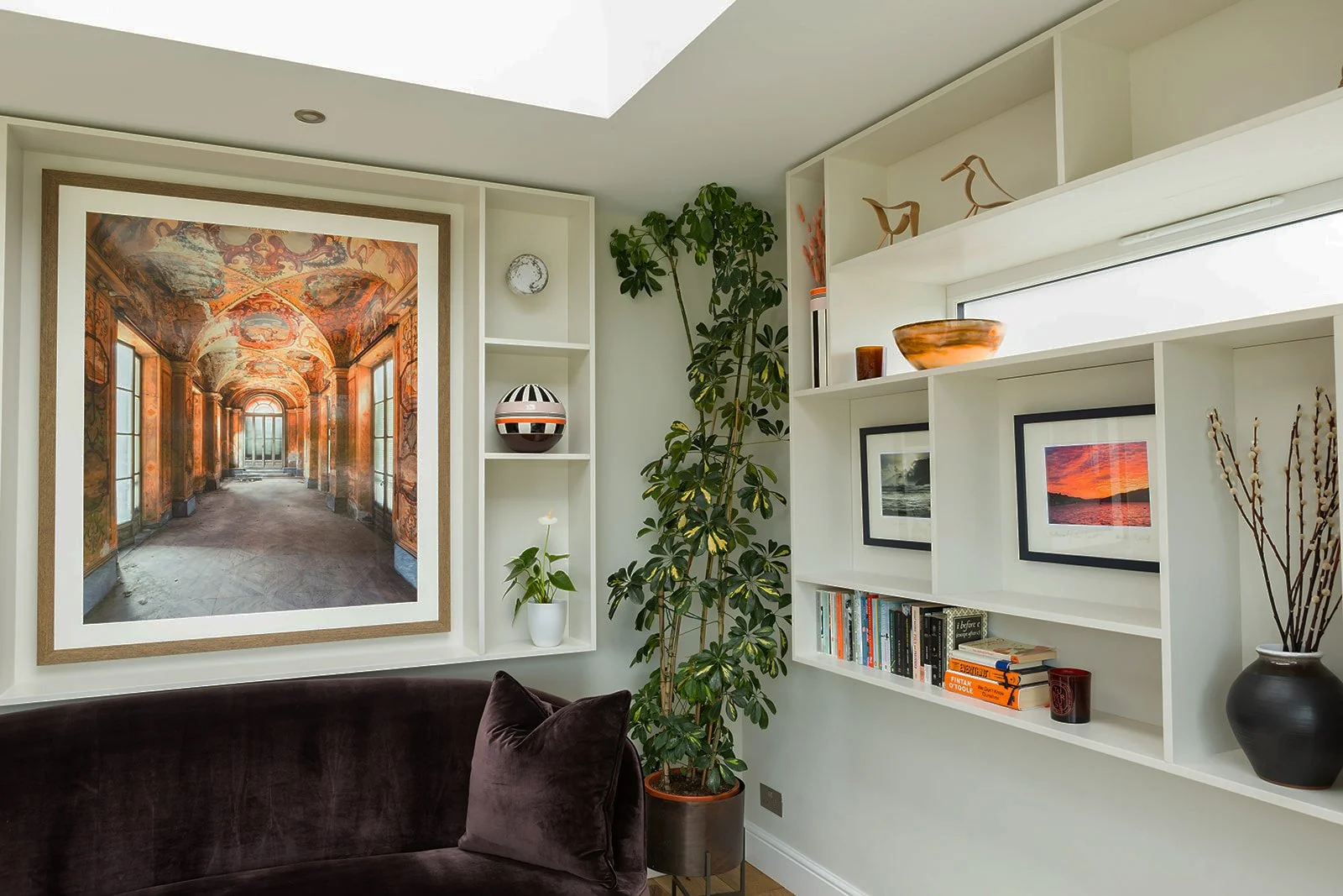 Interior of a living room with a white built-in bookshelf, a tall potted plant, framed sunset photo, framed ocean photo, and decorative objects, with a large wall art piece of an ornate hallway on the wall.