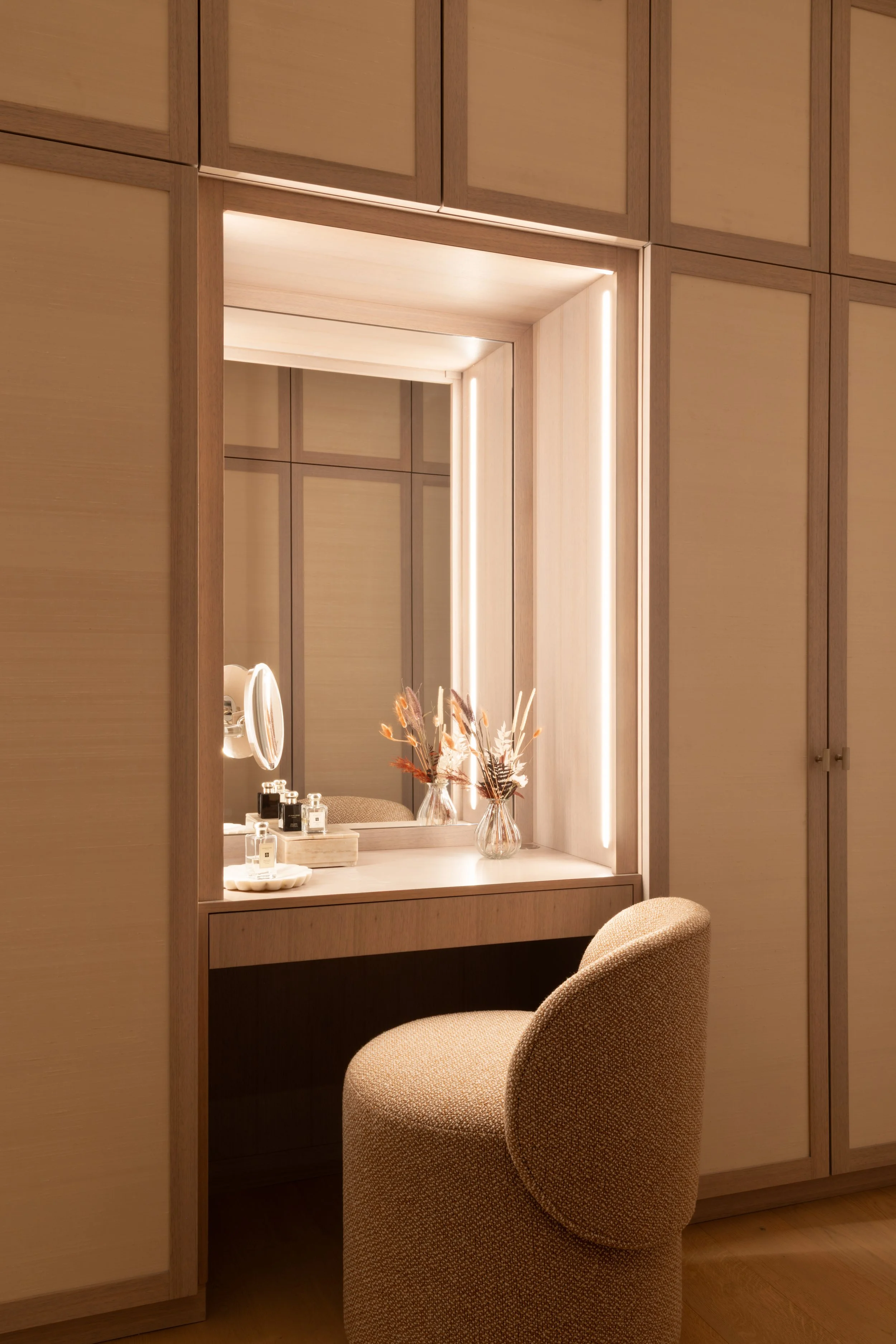 A dressing area with a large mirror framed by vertical light panels, a small table with perfume bottles, and a beige upholstered chair in front of a wooden wall and cabinet.