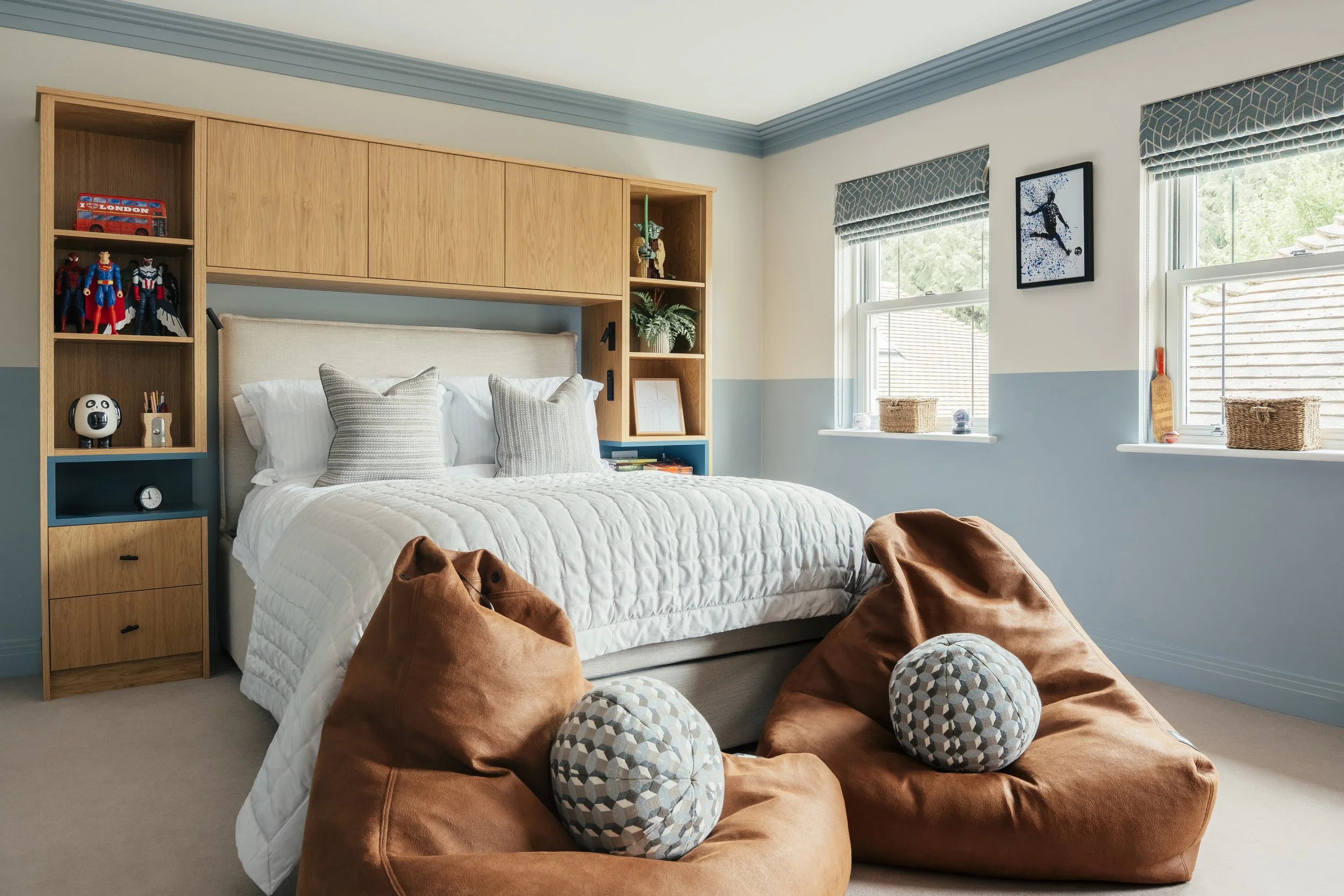 A cozy bedroom with a bed, decorative pillows, a large brown bean bag, and two plush pillows with a honeycomb pattern. The room features a built-in wooden headboard with shelves holding toys, plants, and framed pictures. Two windows with patterned blinds allow natural light, and artwork hangs on the light blue and cream-colored walls.
