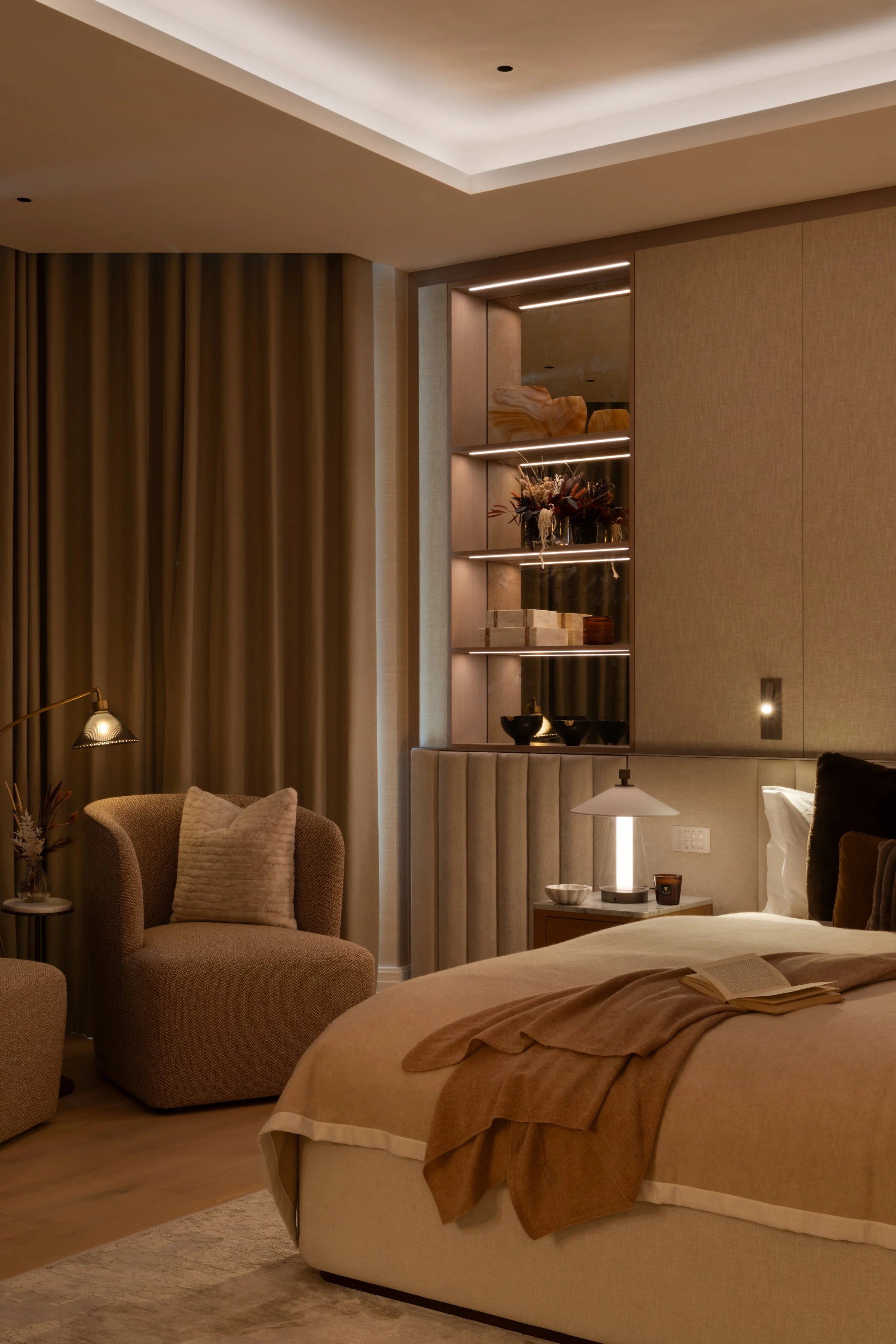 A cozy hotel room with a bed, beige upholstered headboard, and warm lighting. There is a bedside table with a lamp, a book, and decorative items. A comfortable armchair with a pillow and a floor lamp are nearby. The room has beige and gold tones, with built-in shelves displaying a flower arrangement and decorative pieces. Curtains are drawn to one side, revealing soft ceiling lighting.