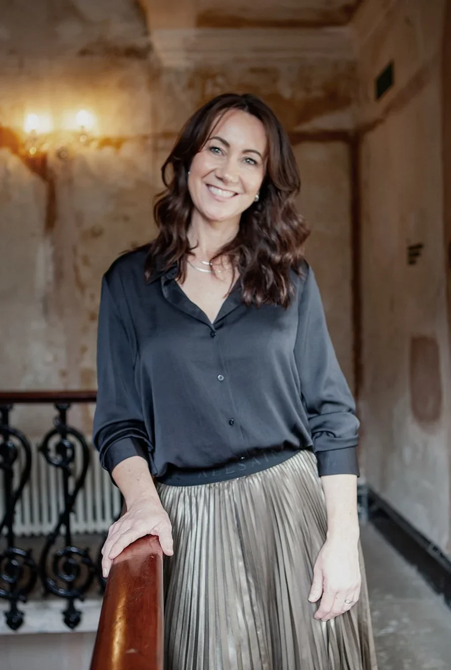 A woman with shoulder-length brown hair, wearing a black blouse and a pleated metallic skirt, standing indoors with one hand on a wooden railing, smiling at the camera.