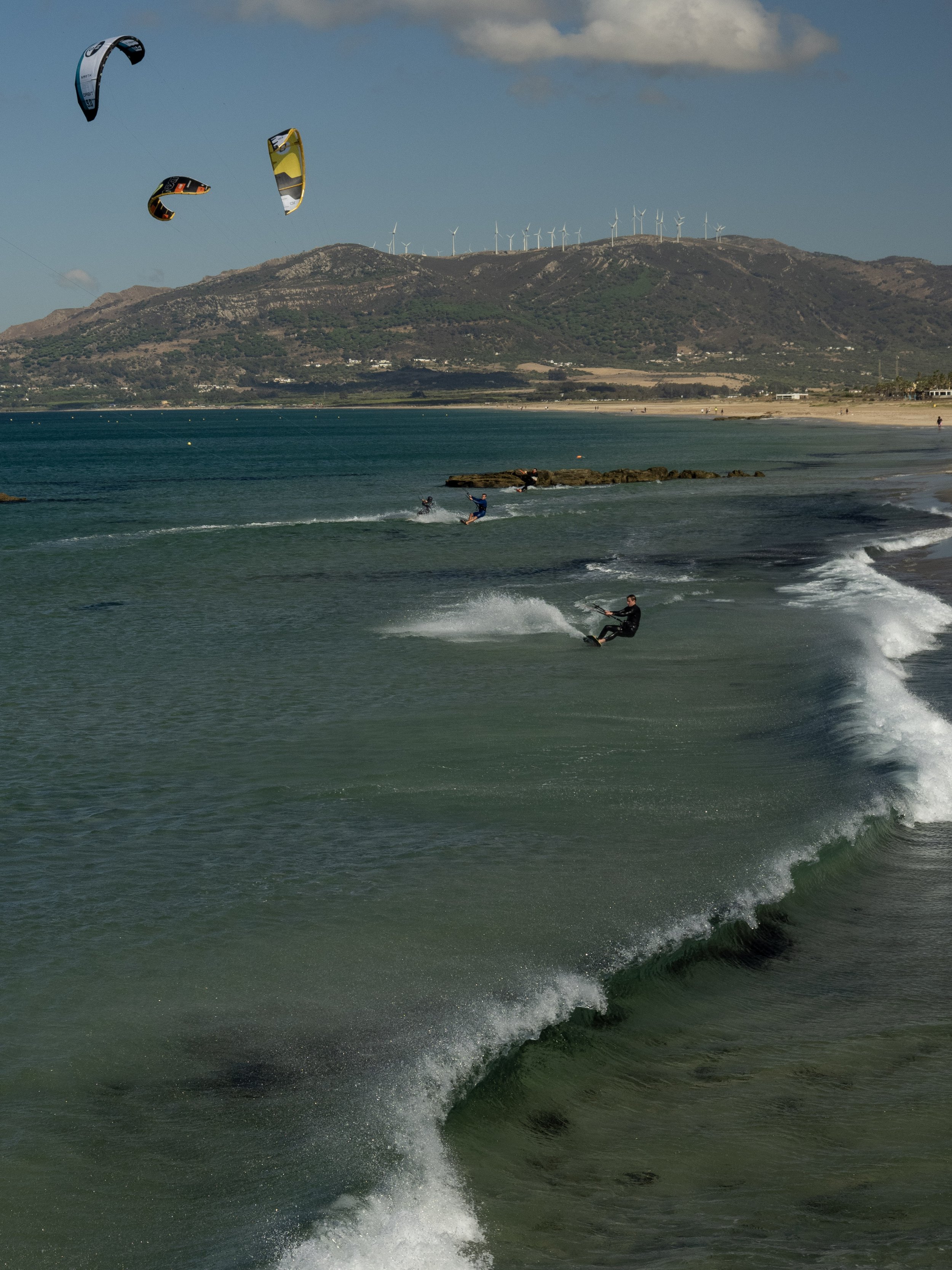 Kitesurfers riding waves near a sandy beach, with wind turbines on the rocky mountain ridge in the background.