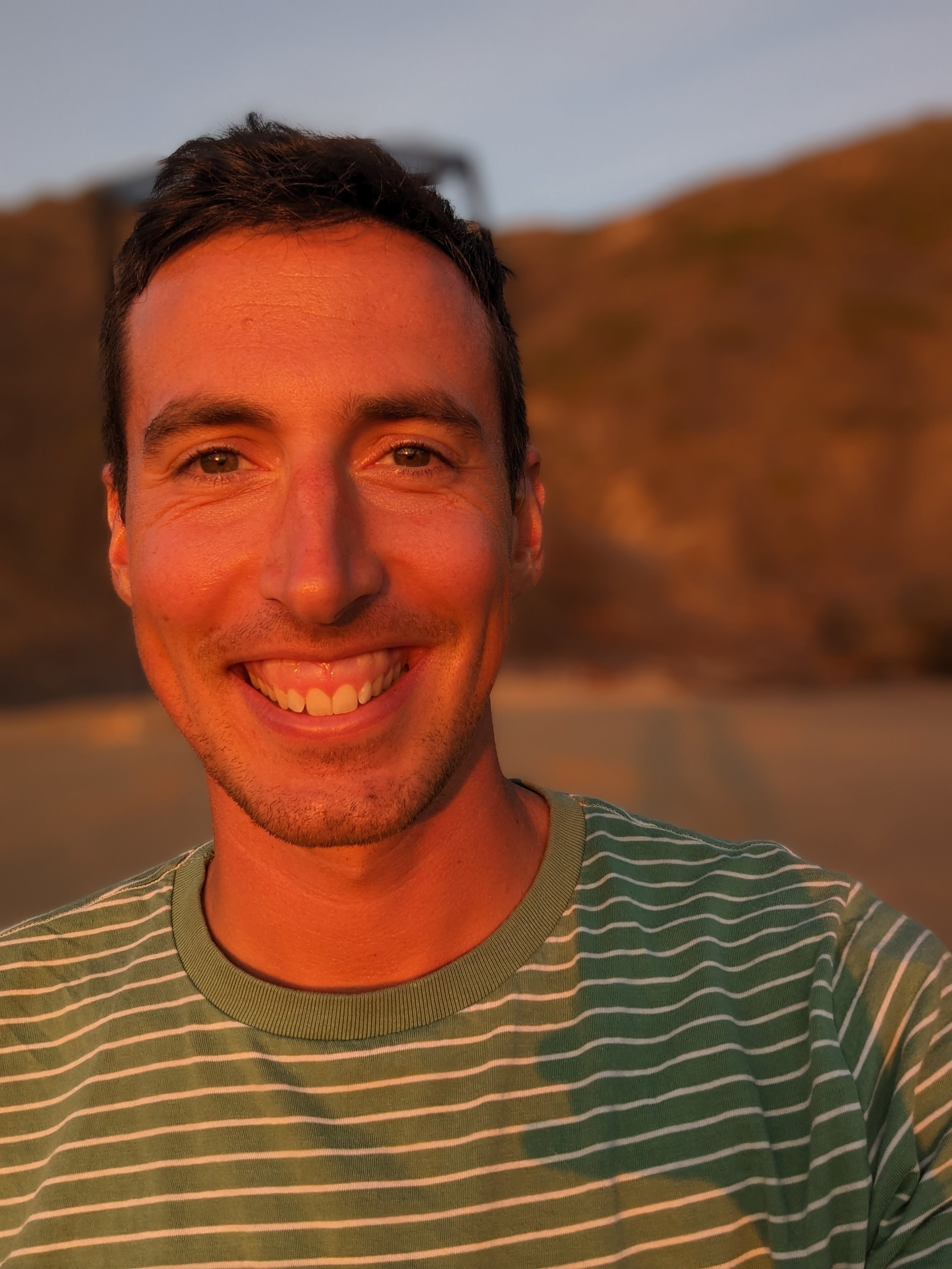 Smiling man in a green and white striped shirt posing outdoors at sunset with blurred mountains in the background