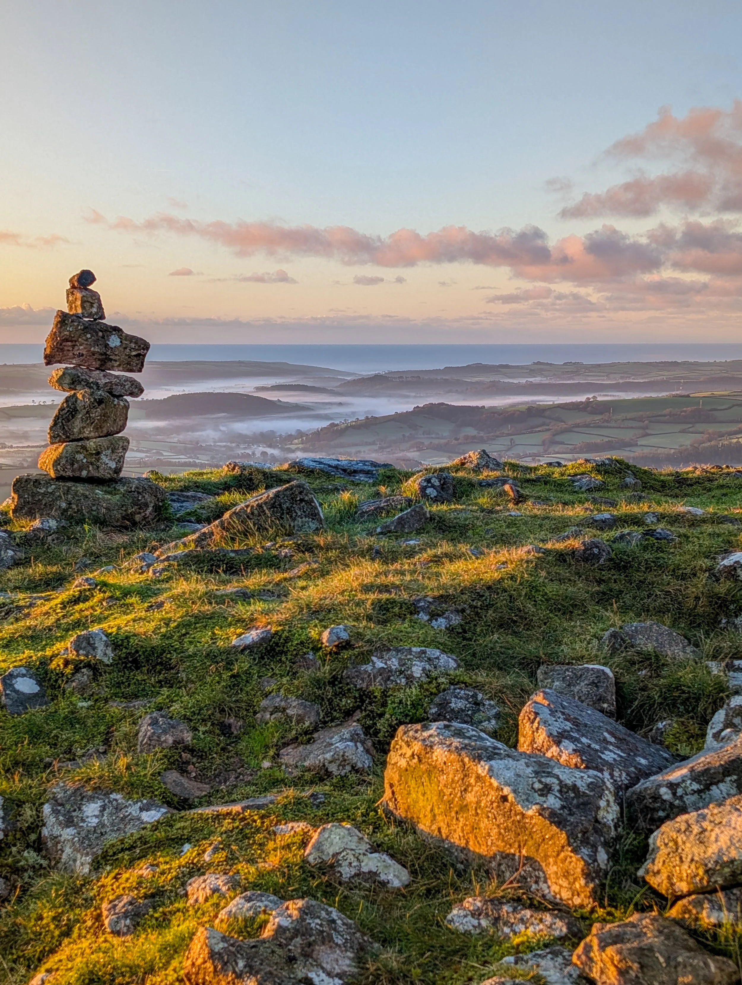 Stacked stone cairn overlooking a misty coastal landscape and green fields at sunset