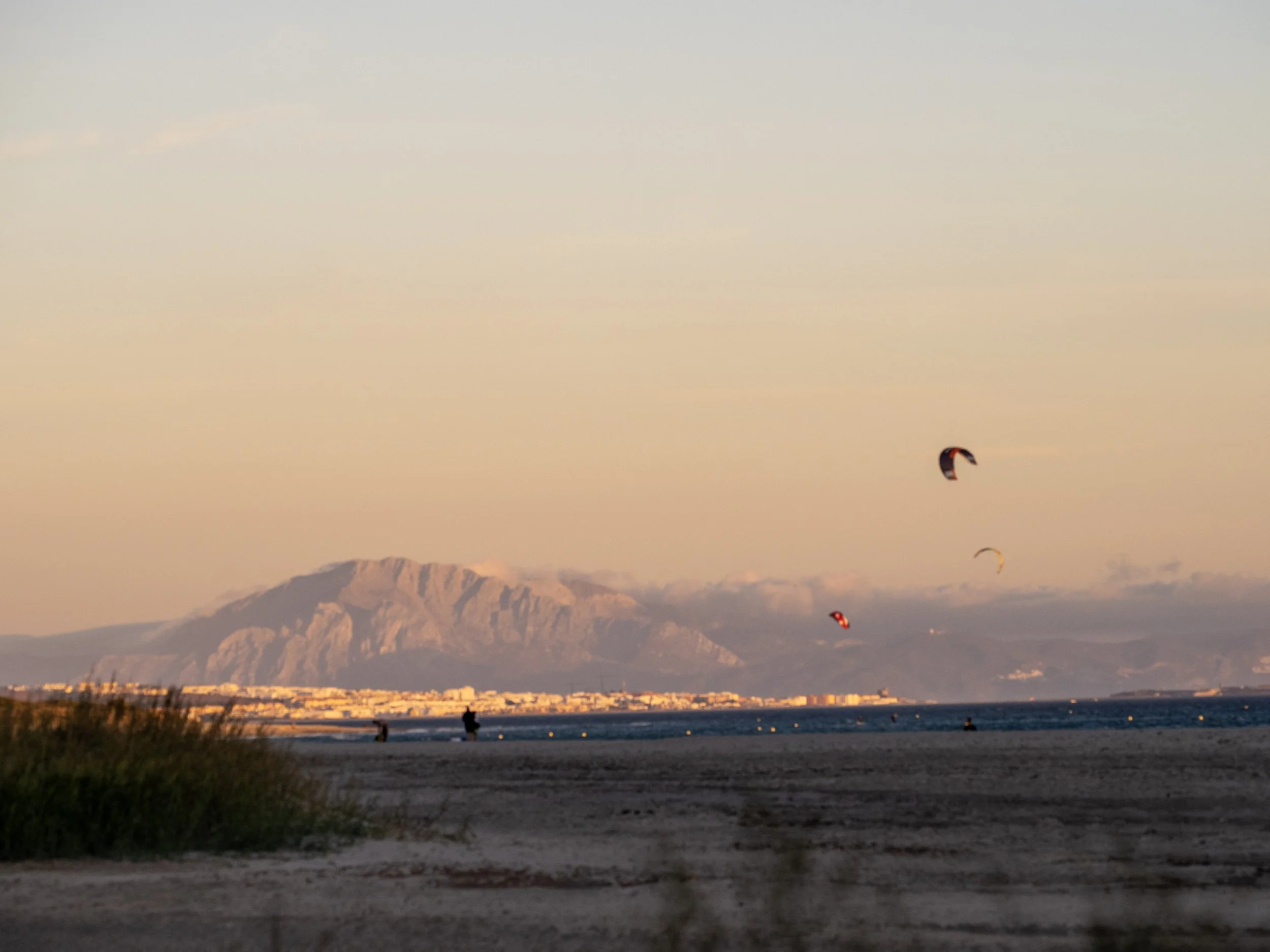Kitesurfers flying over a beach at sunset with a mountain range and coastal city in the background