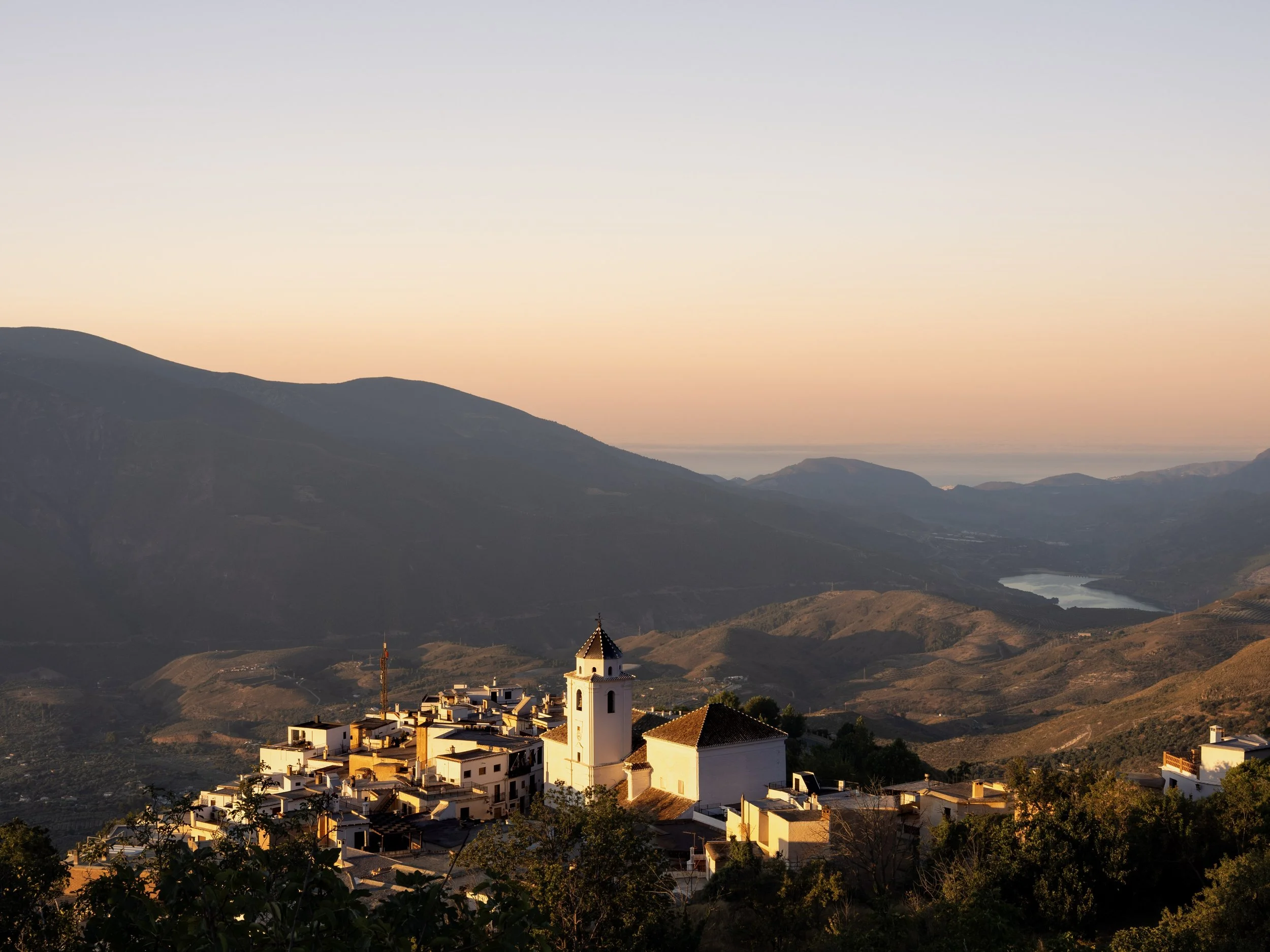 Sunset over a Spanish mountain village with a white church, rugged hills, and a reservoir in the valley