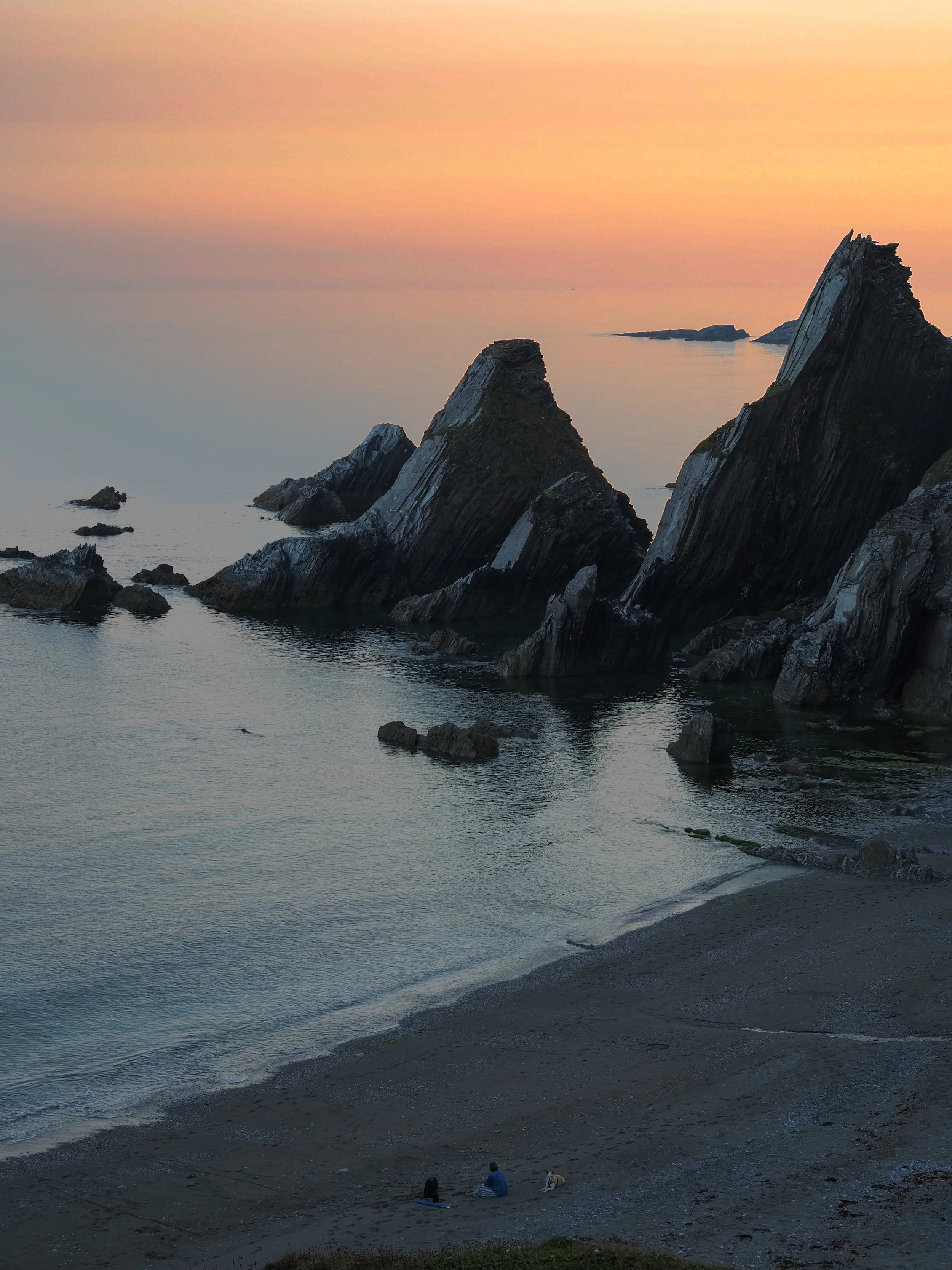 Rugged coastal cliffs and sea stacks above a calm ocean at sunset, with a black pebble beach below