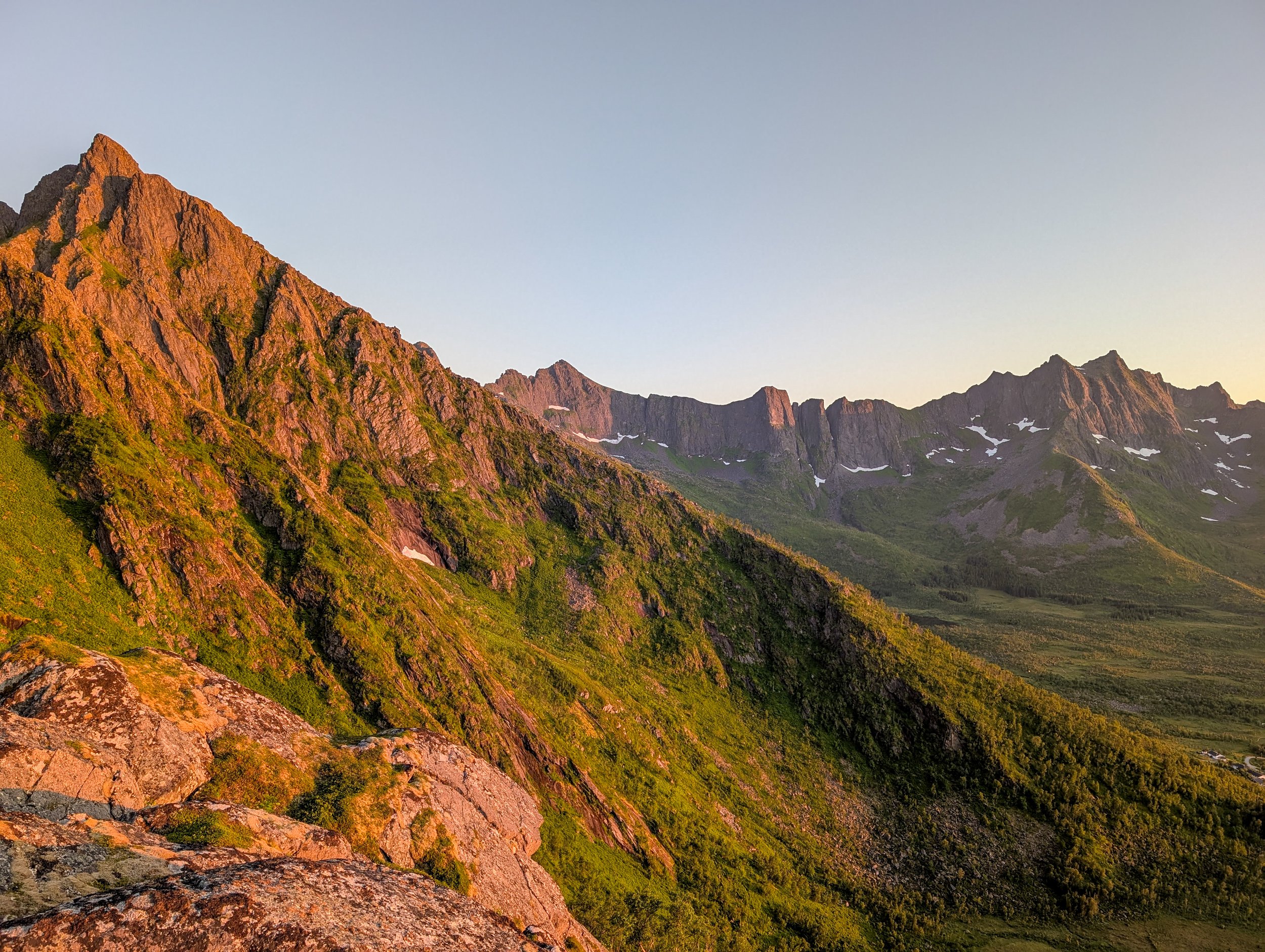 Sunlit rugged mountain range with green slopes, rocky peaks, and patches of snow under a clear sky