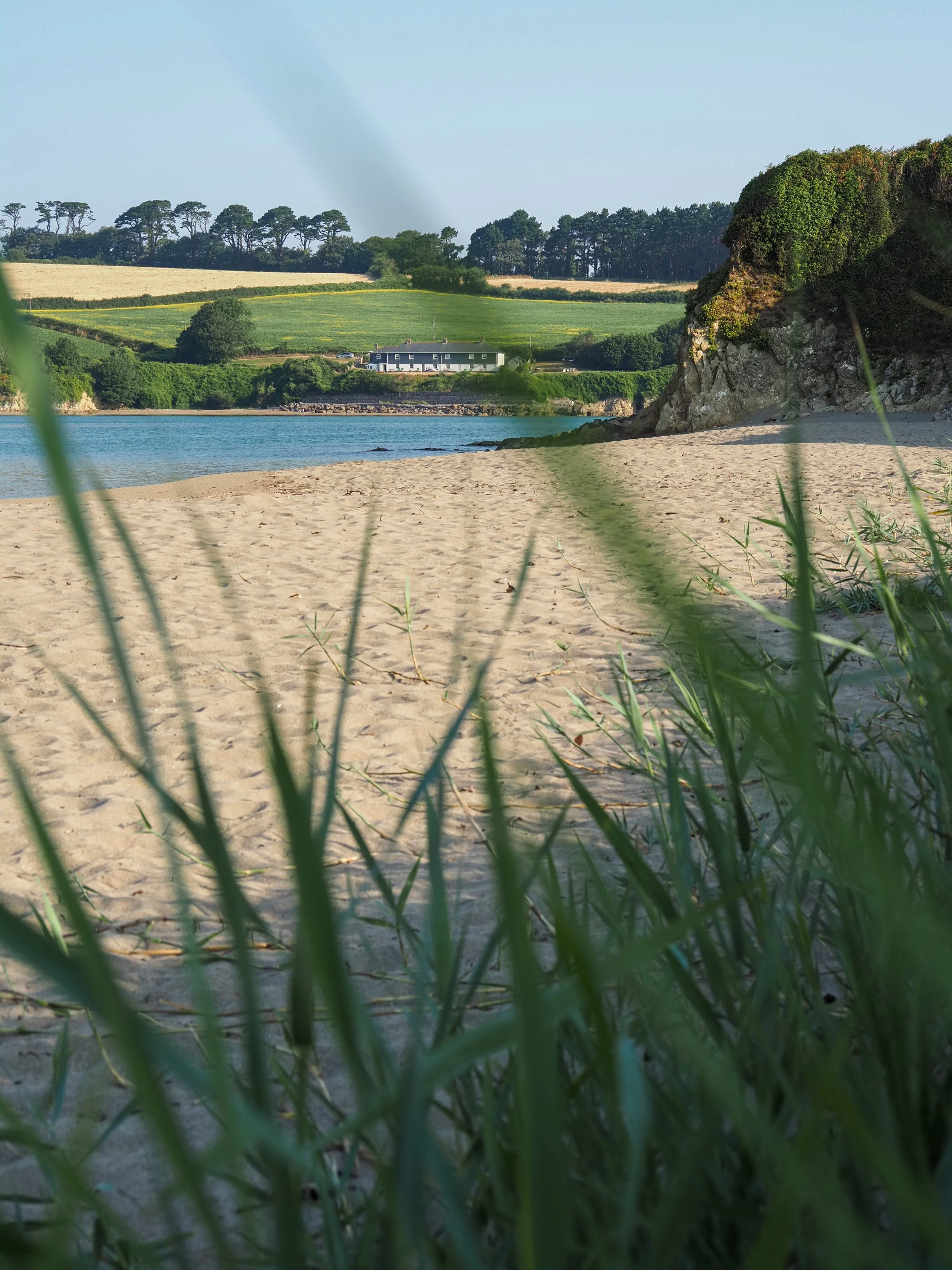 Sandy beach and blue coastal water with rocky cliffs, a farmhouse, green fields, and trees beyond.