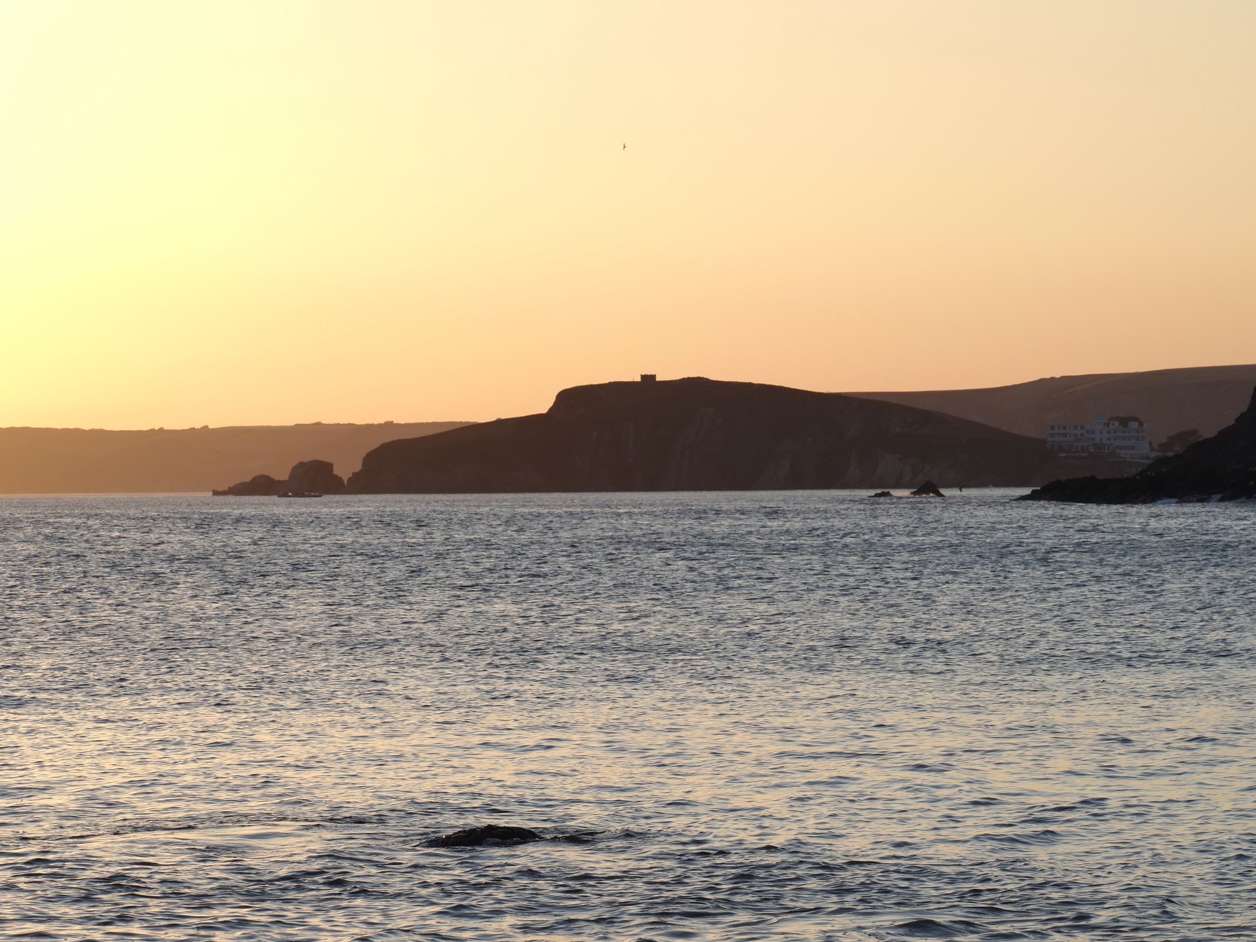 Sunset over a rugged coastal cliff and calm ocean, with a white seaside building on the shoreline.