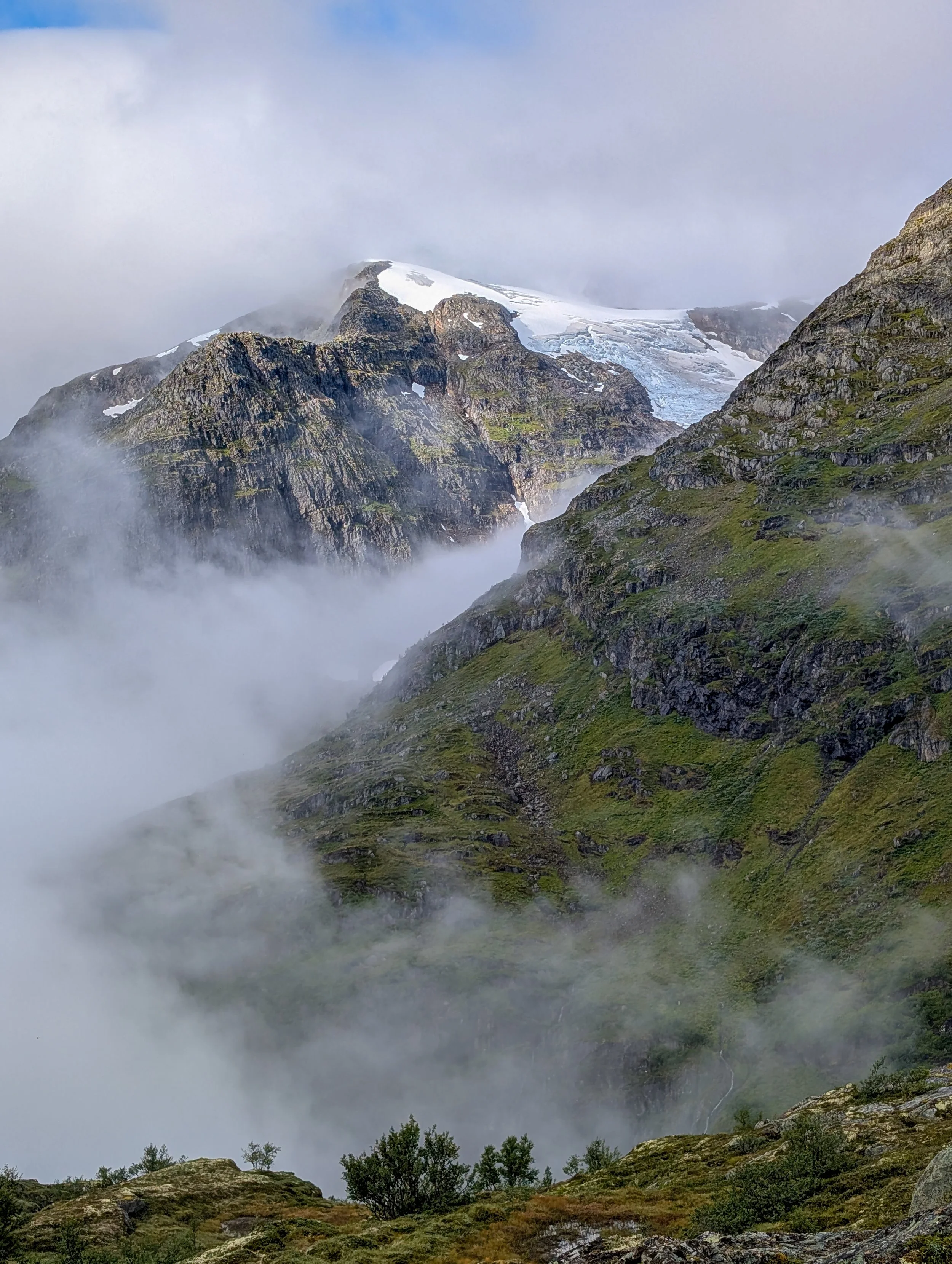 Misty alpine mountain landscape with rugged green cliffs, a glacier, and snow-covered peaks.