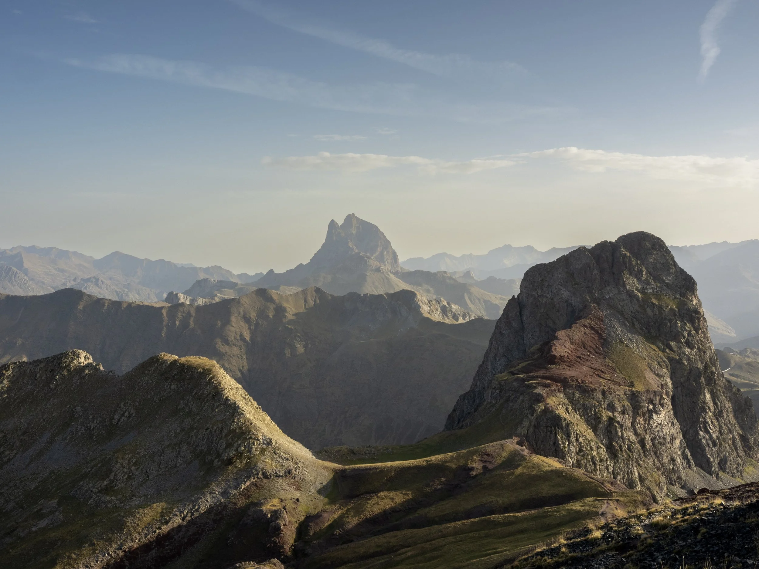 Panoramic view of rugged alpine mountains with jagged peaks and rocky foreground ridges beneath a hazy blue sky