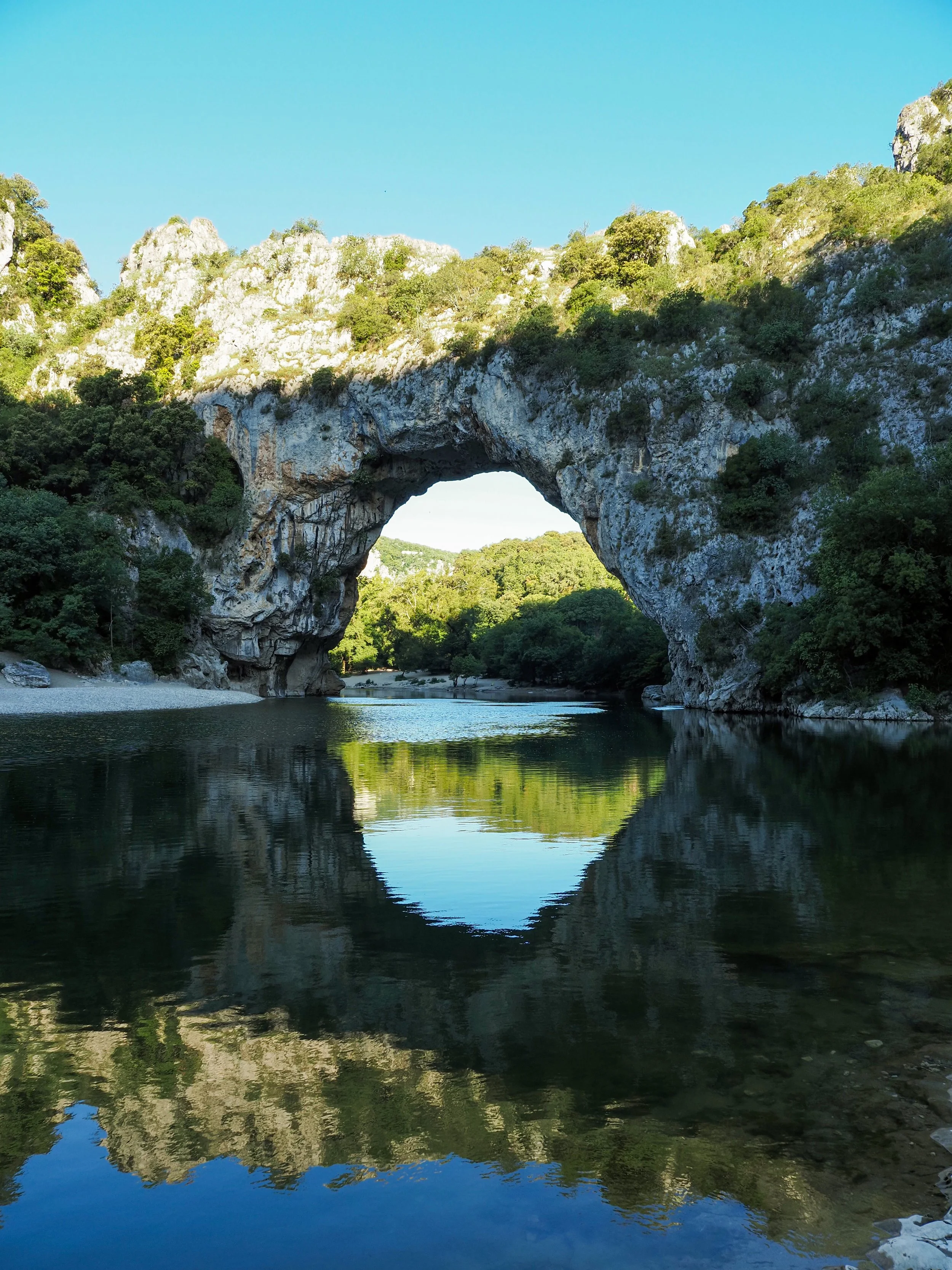 Natural limestone rock arch surrounded by lush greenery over a calm reflective body of water