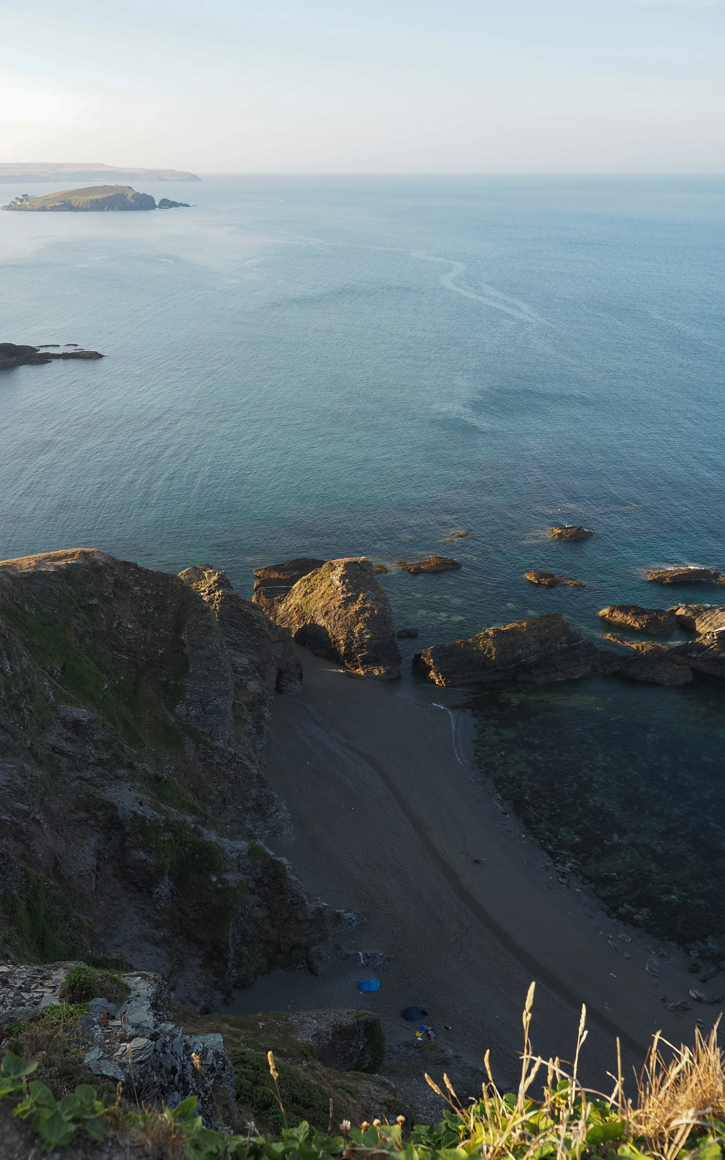 Rocky coastal cliffs and a secluded dark-sand beach overlooking calm blue ocean waters and distant headlands