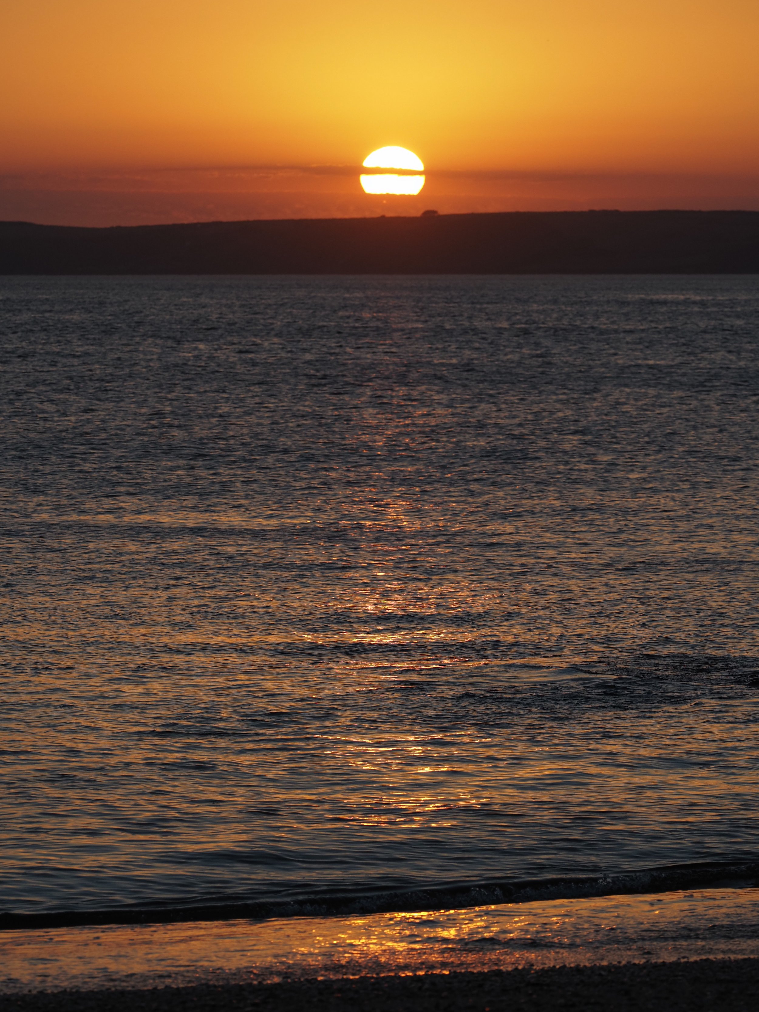 Sunset over the ocean with a glowing sun, silhouetted horizon, and reflections on rippling water