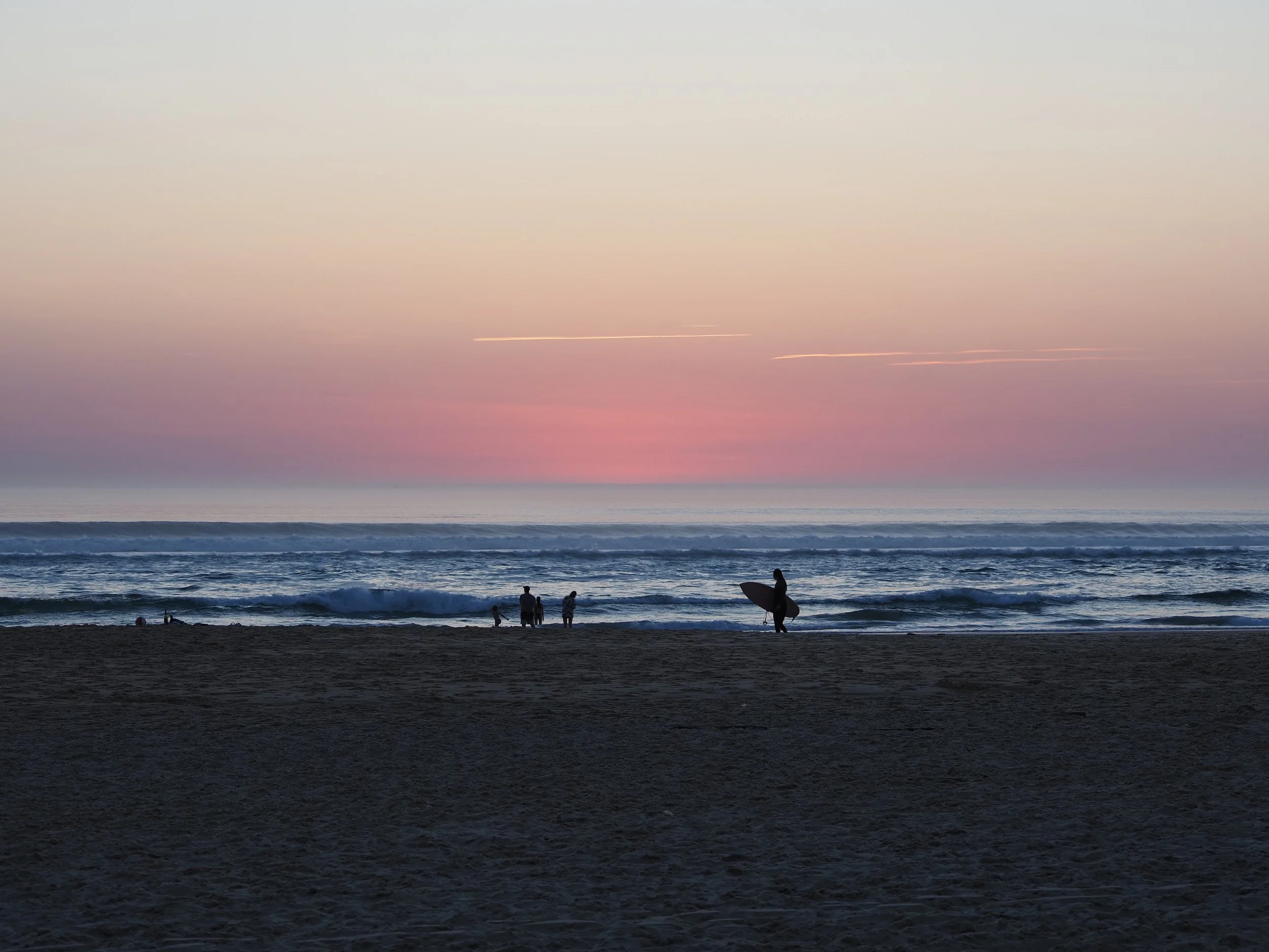 Surfer carrying a surfboard along a beach at sunset with ocean waves and silhouetted beachgoers